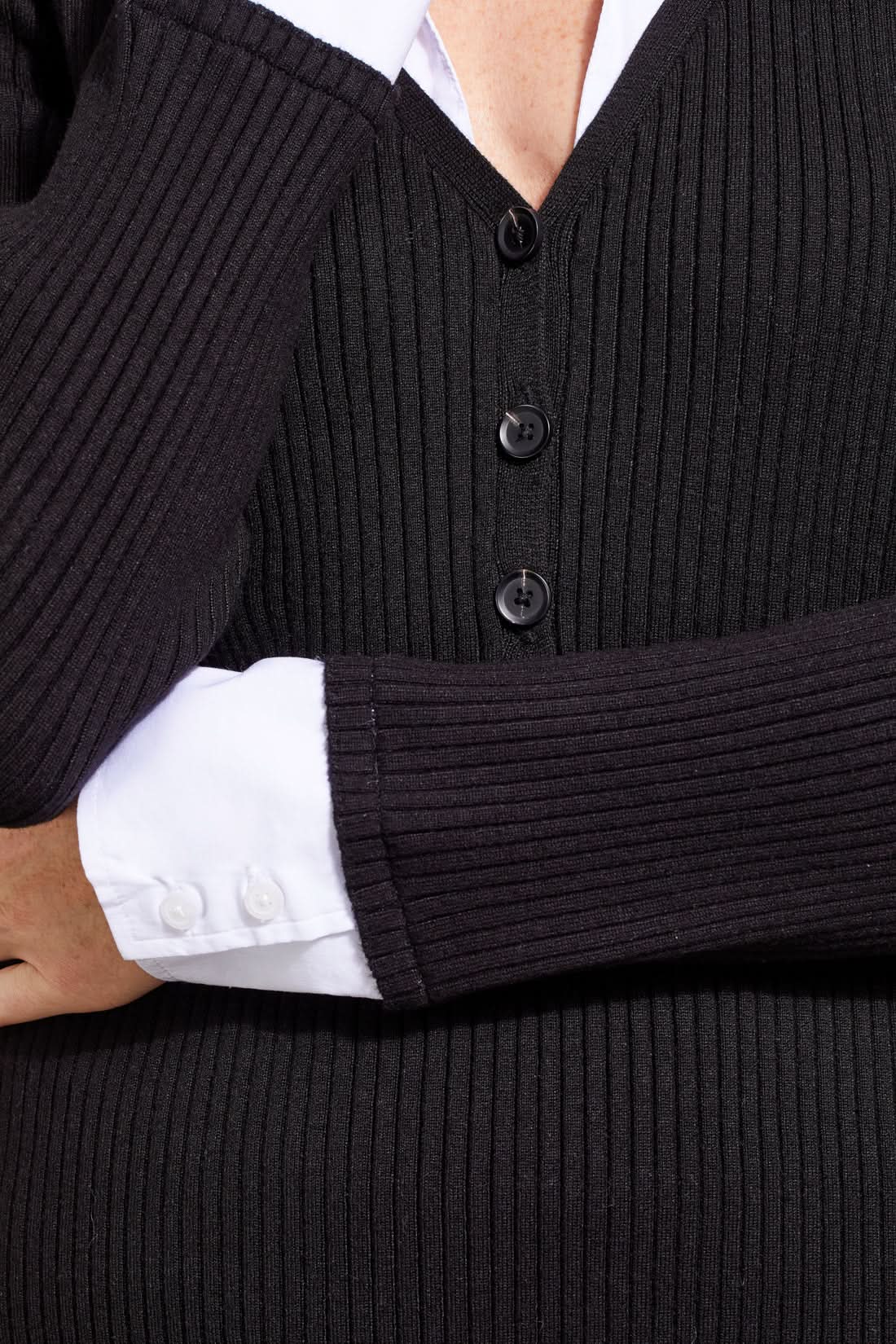 A person with shoulder-length hair is wearing a black ribbed cardigan with buttons over a white collared shirt, creating a fashionable look. The Tribal Henley Fooler Sweater adds an extra touch of sophistication, crafted from high-quality materials for enduring style.