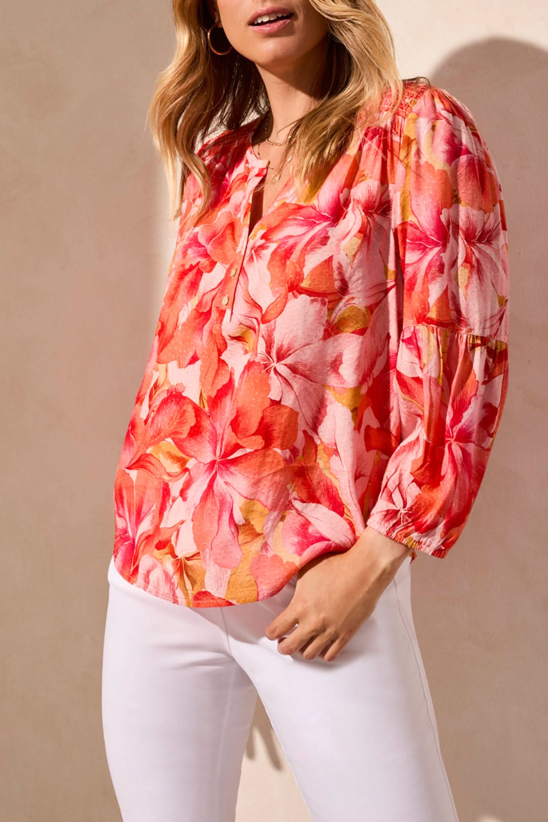 Someone in the Tribal 3/4 Sleeve Blouse with Smocked Yoke, featuring a vibrant floral pattern in pink and orange, paired with white pants against a neutral background.
