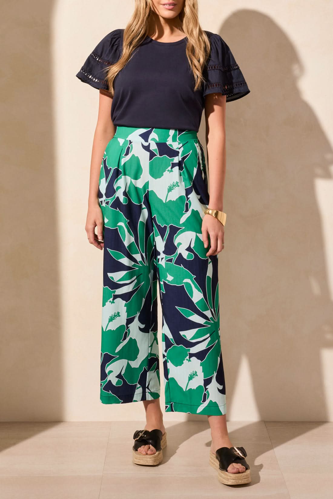 Against a neutral backdrop, an individual wears a navy top matched with Tribal's Pull On Ankle Pant with Pleats in a green-and-navy floral design. This stylish and versatile outfit blends functionality and effortless style, making it ideal for any occasion.