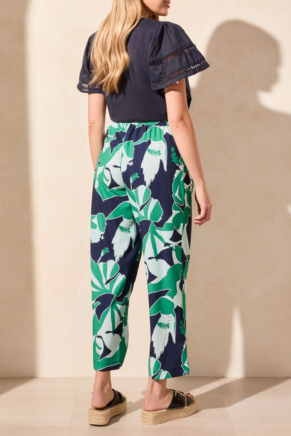 Against a neutral backdrop, an individual wears a navy top matched with Tribal's Pull On Ankle Pant with Pleats in a green-and-navy floral design. This stylish and versatile outfit blends functionality and effortless style, making it ideal for any occasion.
