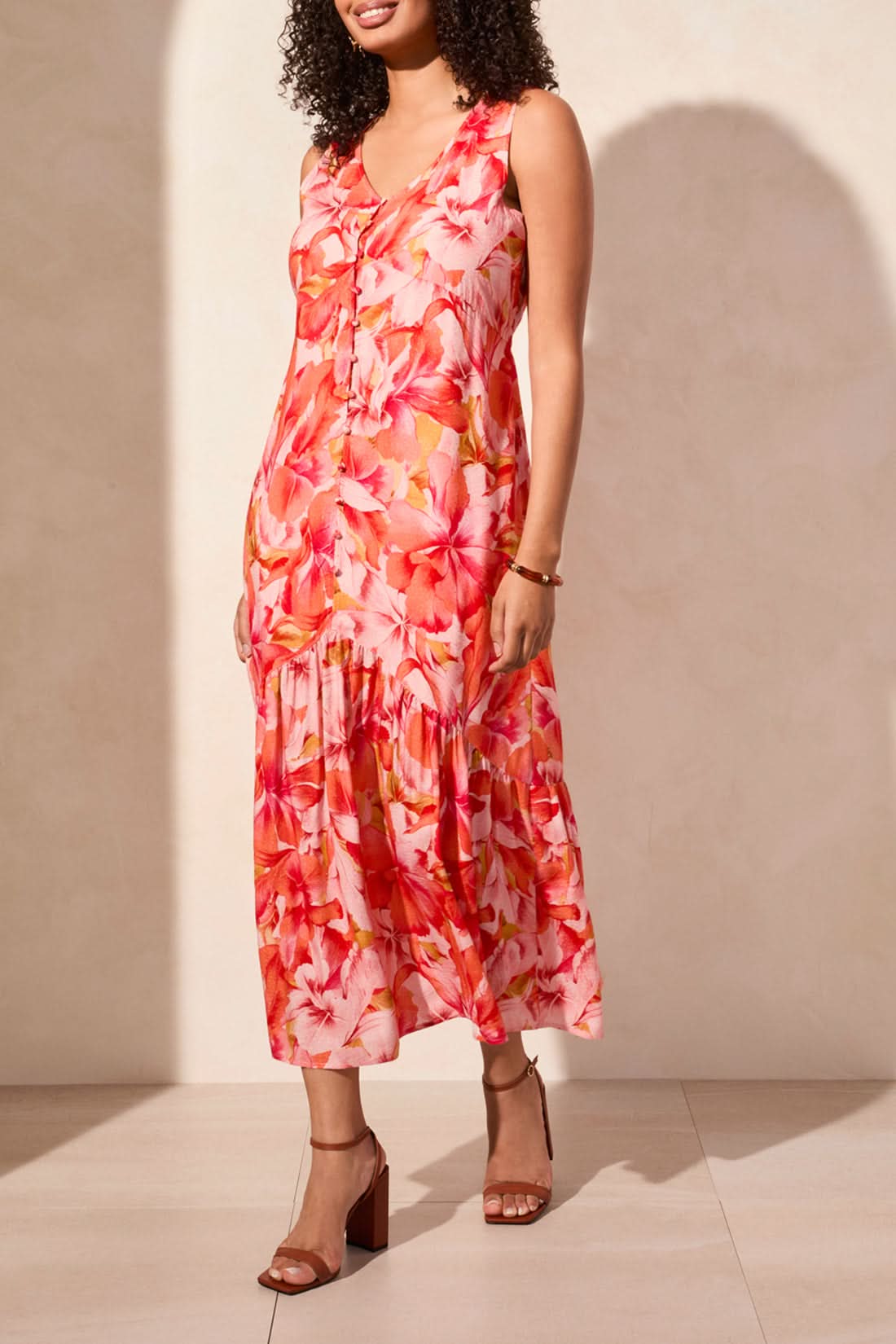 In a Tribal Sleeveless Maxi Dress with Lining, featuring a pink, orange, and white floral pattern, a woman exudes feminine charm against a plain background.