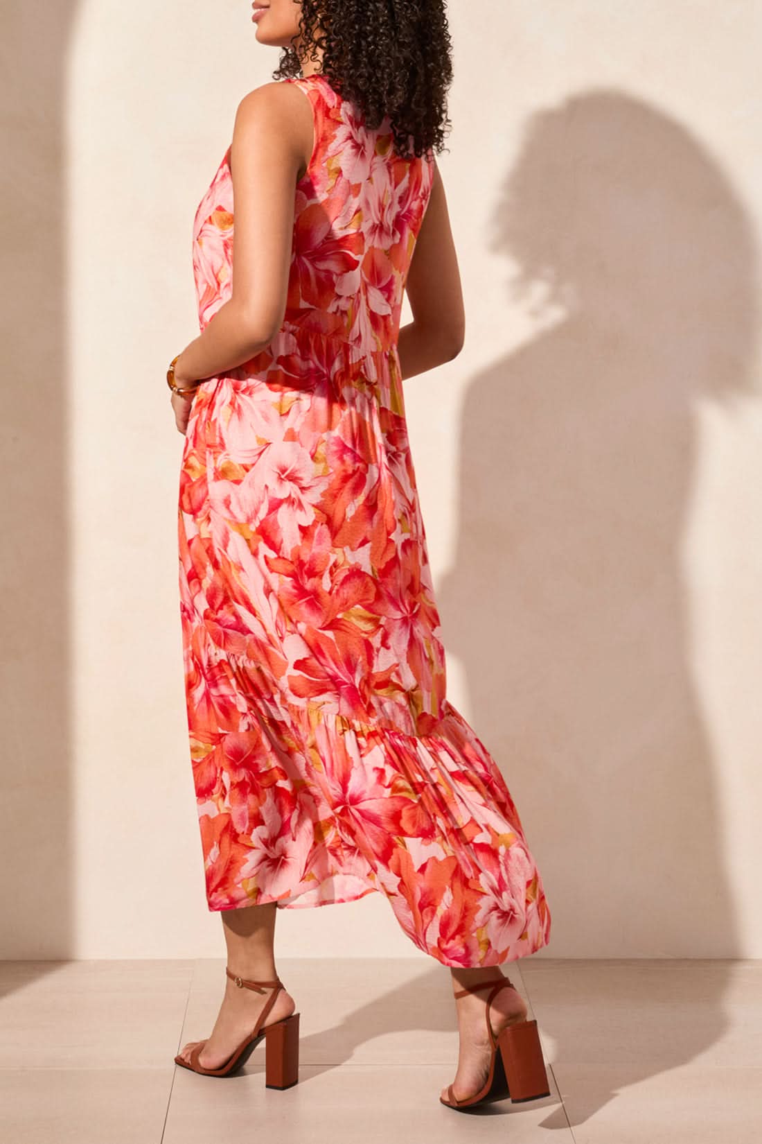 In a Tribal Sleeveless Maxi Dress with Lining, featuring a pink, orange, and white floral pattern, a woman exudes feminine charm against a plain background.