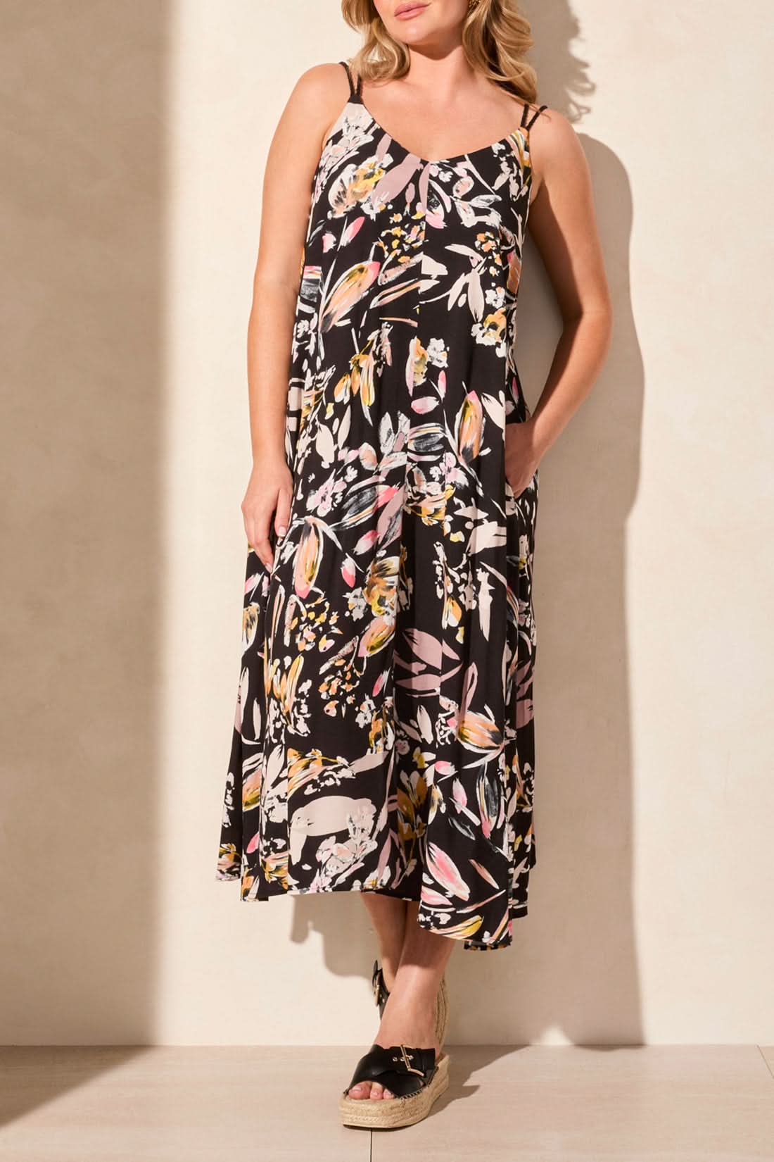 A person stands against a light wall wearing the Tribal brand LINED MAXI DRESS WITH ADJUSTABLE CORDS. The sleeveless black floral dress features a V-neckline and offers elegance and comfort for sunny days, with hands in pockets and paired with wedge sandals.