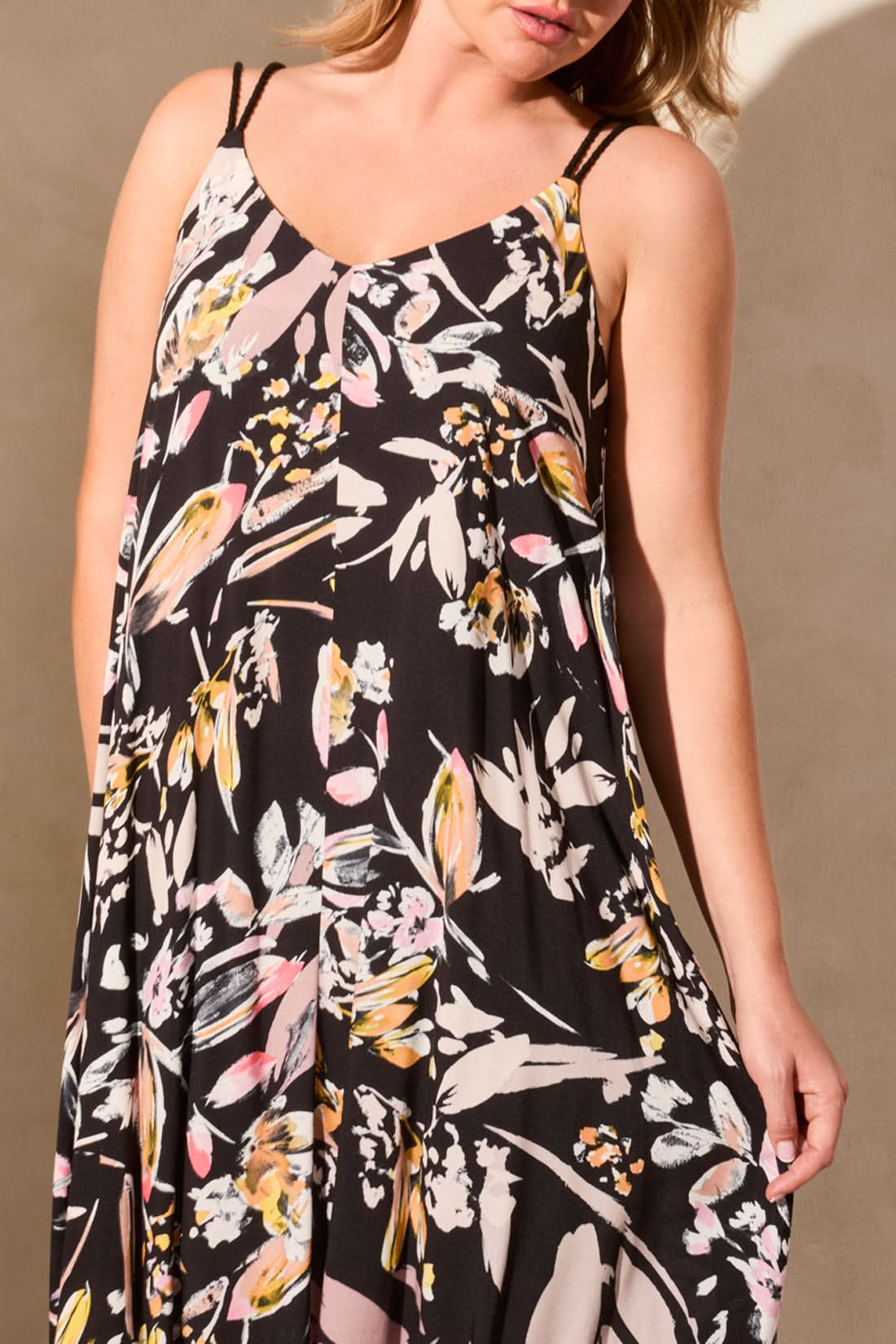 A person stands against a light wall wearing the Tribal brand LINED MAXI DRESS WITH ADJUSTABLE CORDS. The sleeveless black floral dress features a V-neckline and offers elegance and comfort for sunny days, with hands in pockets and paired with wedge sandals.