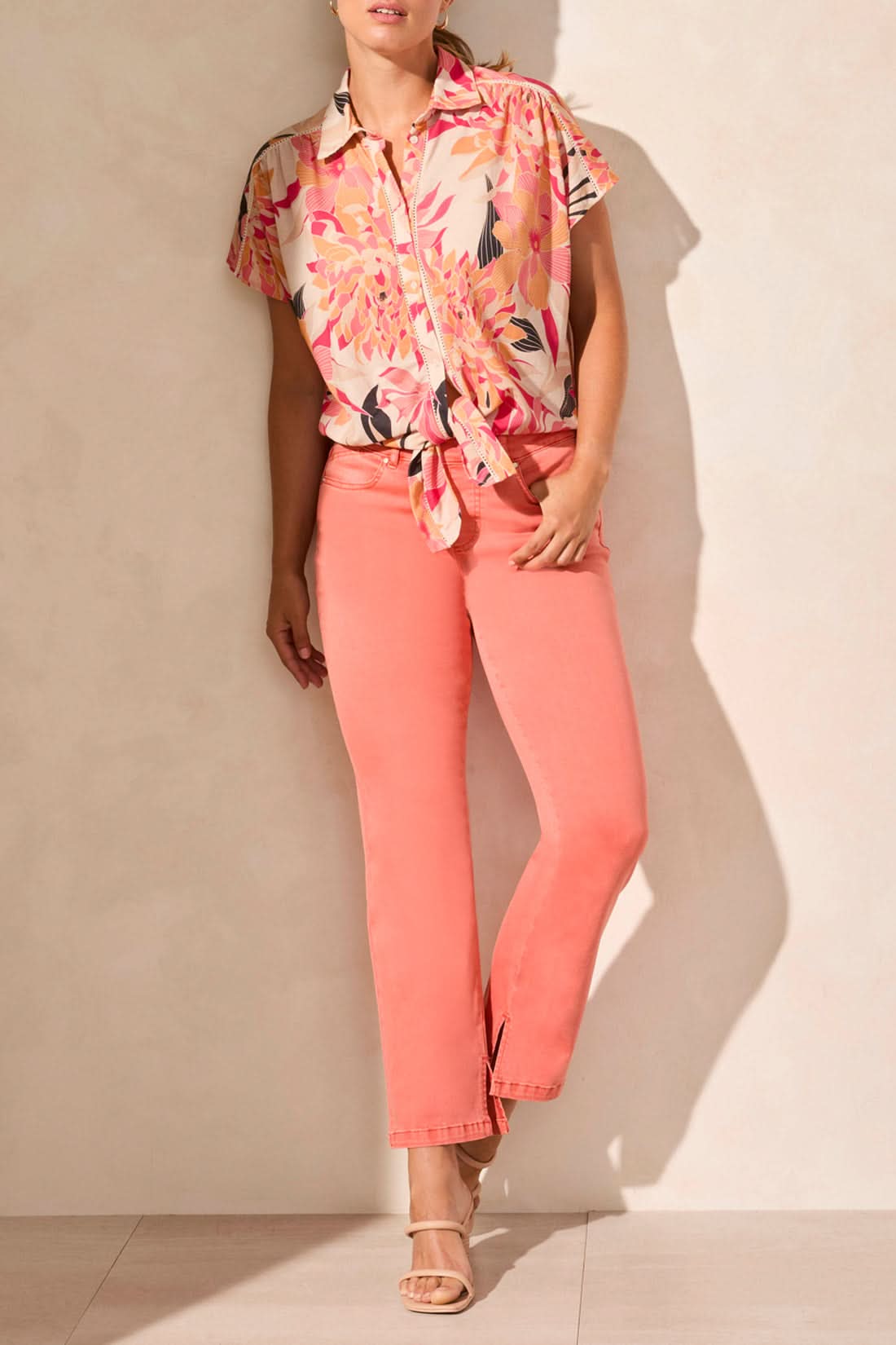 A person in a Tribal short sleeve top featuring buttons and a tie front, paired with coral pants and beige heeled sandals, stands on a light floor, exemplifying the ideal summer look.
