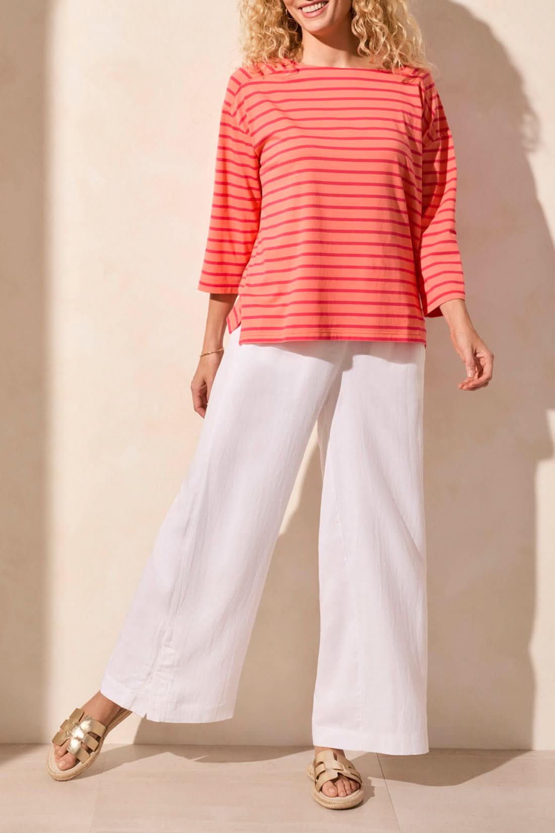 A person wearing a stylish Tribal 3/4 Sleeve Boat Neck Top in coral and pink stripes paired with white pants stands against a beige background.