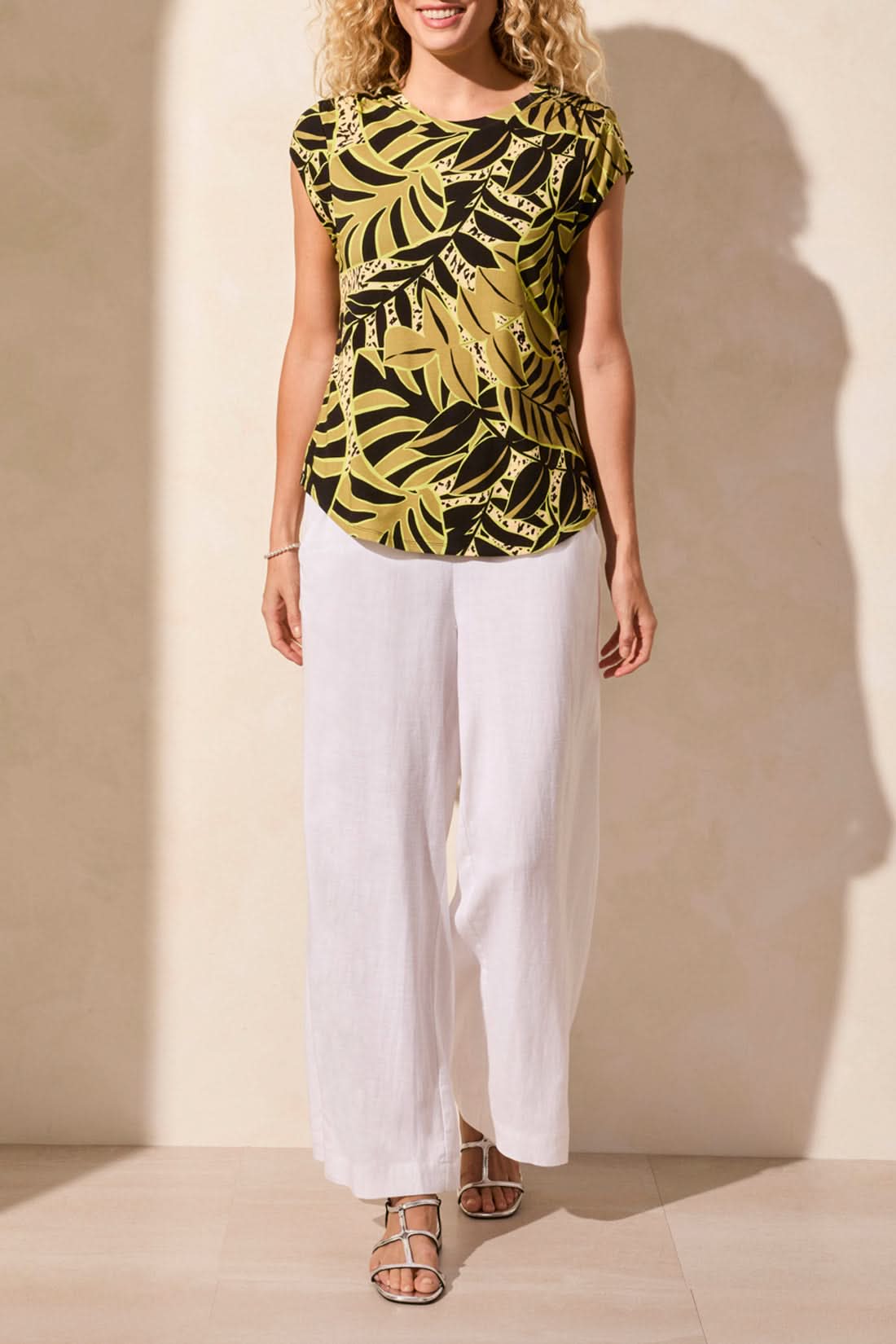 A woman models Tribal's CAP SLEEVE TOP WITH SHIRRING, featuring a green and black tropical leaf print, paired with white pants against a neutral backdrop.
