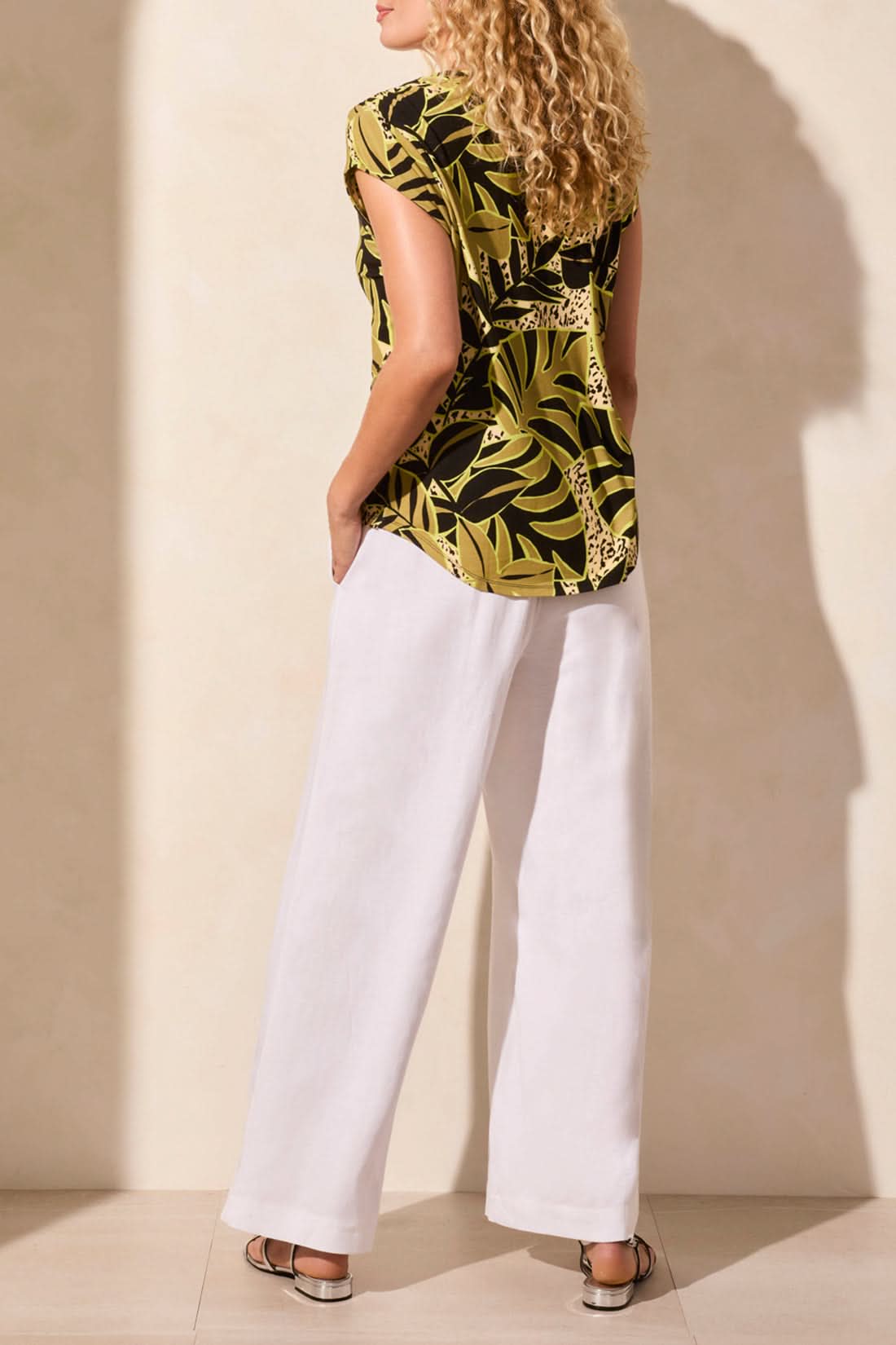 A woman models Tribal's CAP SLEEVE TOP WITH SHIRRING, featuring a green and black tropical leaf print, paired with white pants against a neutral backdrop.
