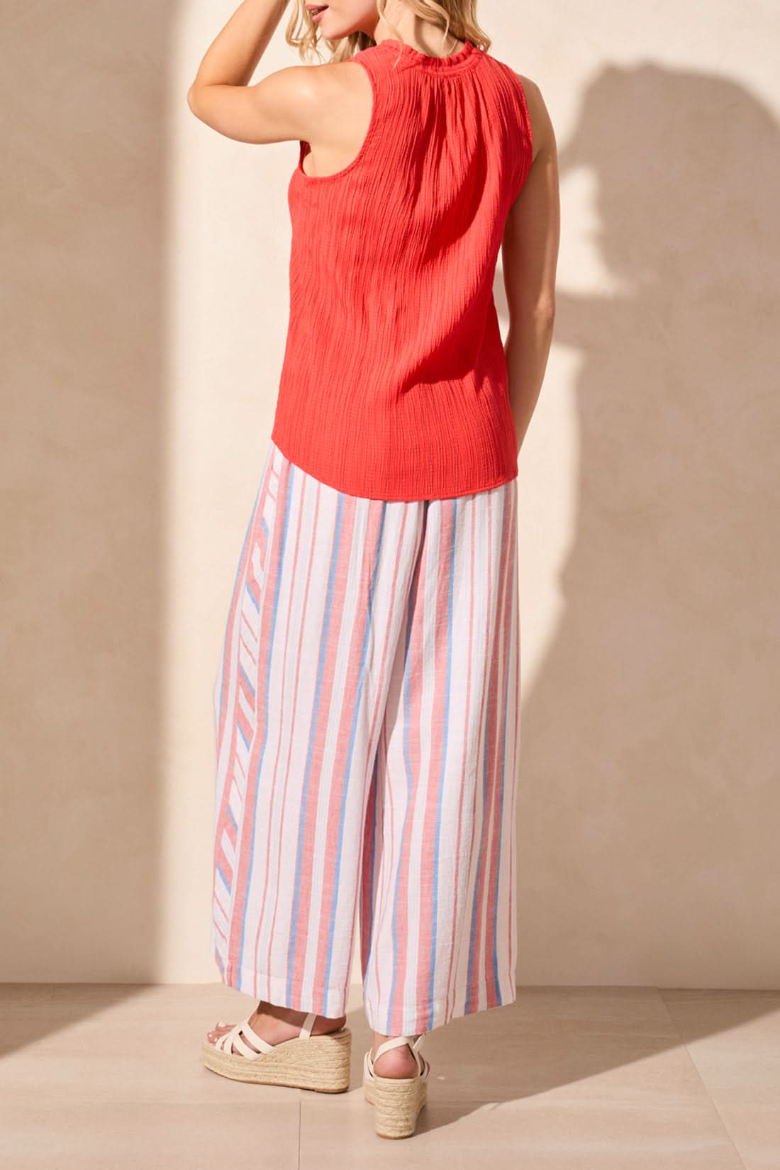 A woman wears the Tribal SLEEVLESS BLOUSE WITH BUTTON PLACKET, featuring a textured red finish, paired with relaxed striped pants as she stands against a light-colored wall.
