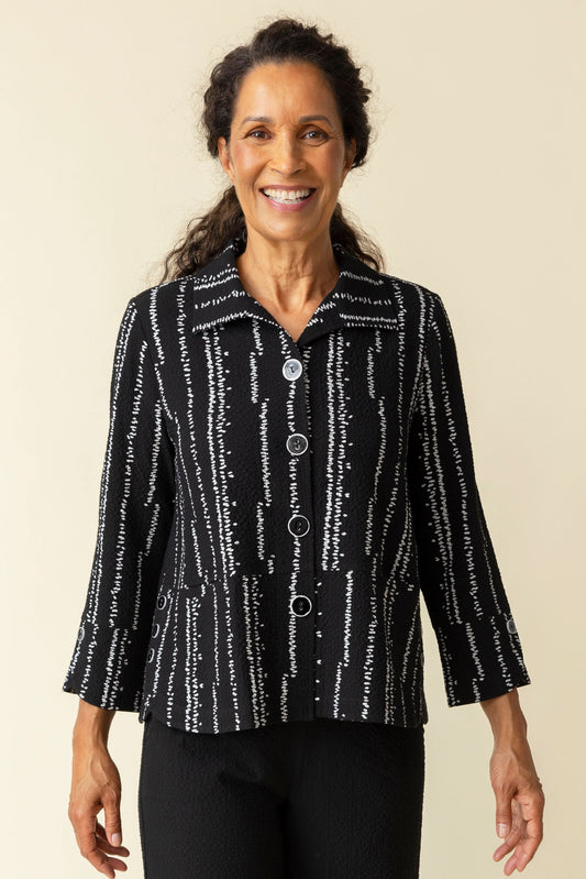A woman smiles in a Habitat Tulip Hem Swing Jacket, featuring black and white patterns, expert tailoring, and three-quarter sleeves, paired with black pants against a plain beige background.