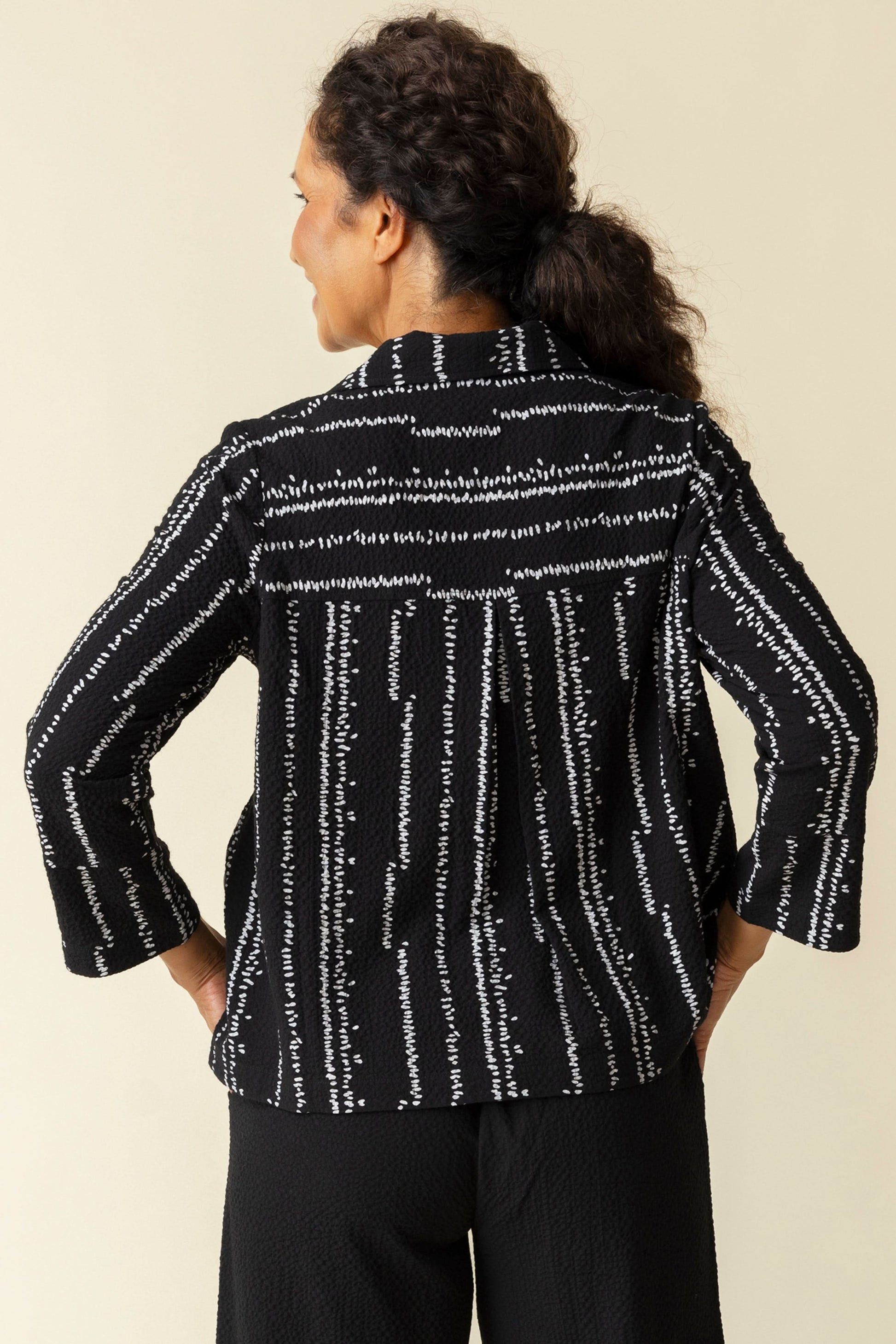 A woman smiles in a Habitat Tulip Hem Swing Jacket, featuring black and white patterns, expert tailoring, and three-quarter sleeves, paired with black pants against a plain beige background.