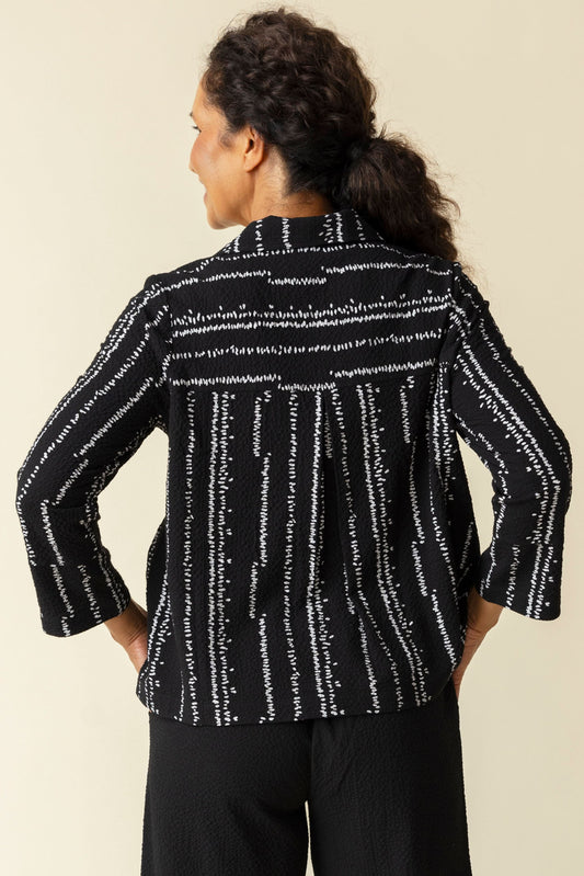 A woman smiles in a Habitat Tulip Hem Swing Jacket, featuring black and white patterns, expert tailoring, and three-quarter sleeves, paired with black pants against a plain beige background.