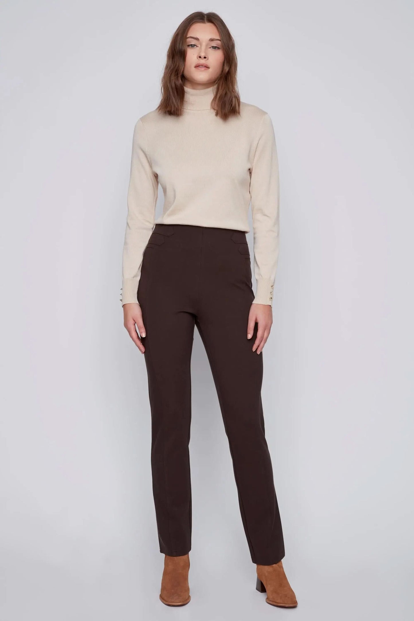 A person with long hair stands against a plain background, showcasing effortless style in a beige turtleneck sweater, dark brown high-waisted CoCo Y Club Pull On Pant, and brown ankle boots.