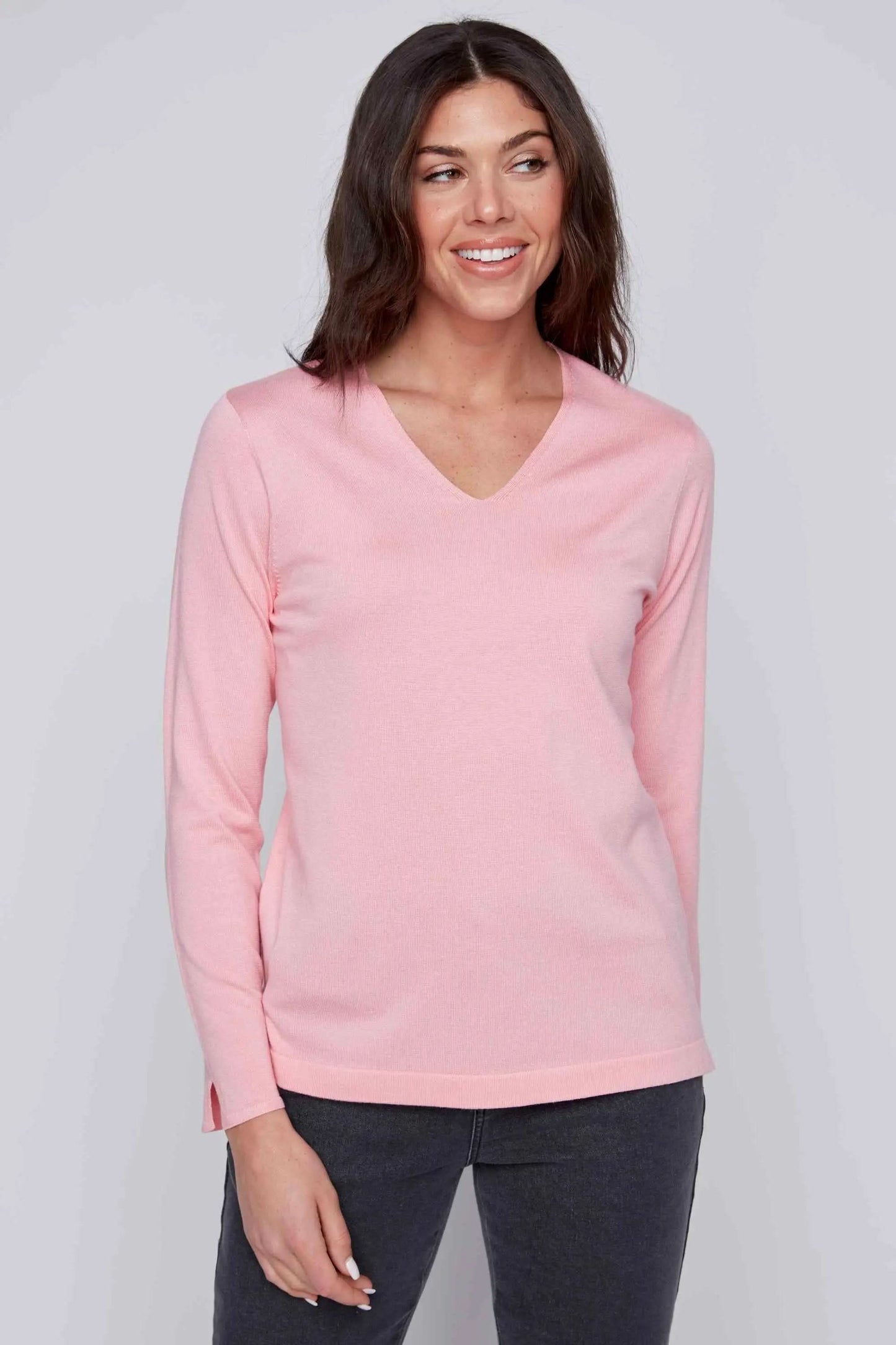 A woman in a CoCo Y Club V Neck Pull Over (light pink, soft material) and dark pants stands against a plain gray background, smiling slightly.
