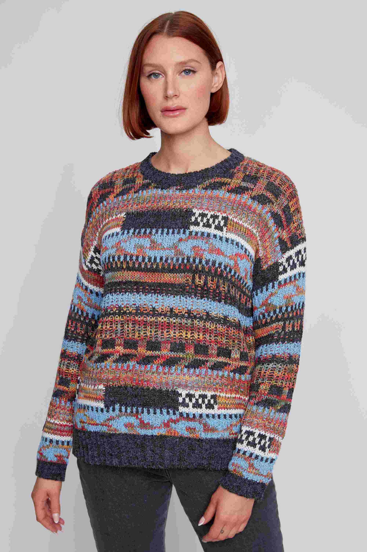 A person with short red hair is wearing a CoCo Y Club Crew Neck Long Sleeve Sweater, featuring a multi-colored design and made of super soft material, paired with dark pants. They are standing against a plain gray background.
