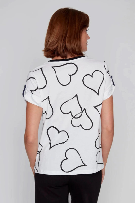 A person stands against a plain background, wearing a CoCo Y Club T-Shirt of Hearts featuring black-outlined hearts on white, paired with classic black pants.