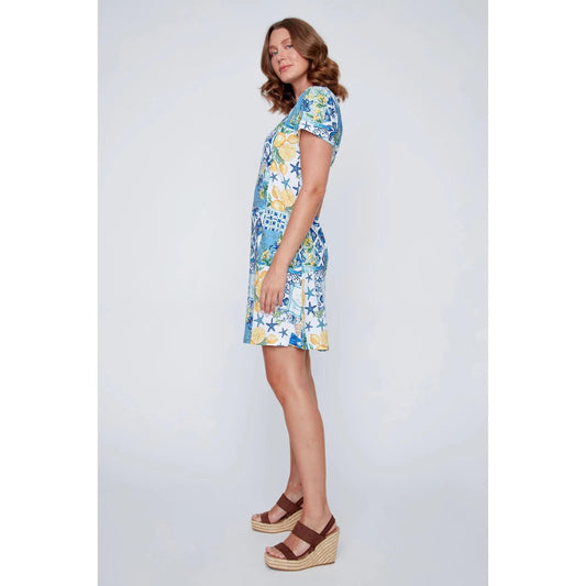 A woman stands smiling in the CoCo Y Club ‘Amalfi’ Lace Detail Dress, a short sleeve style featuring blue and yellow Greek-inspired tile and lemon motifs on a white background.
