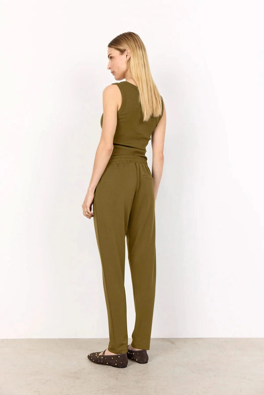 A woman stands against a white wall wearing Soya Concepts Dark Olive Sweatpants with an adjustable waist, hands in pockets, paired with a sleeveless top and brown studded flats.