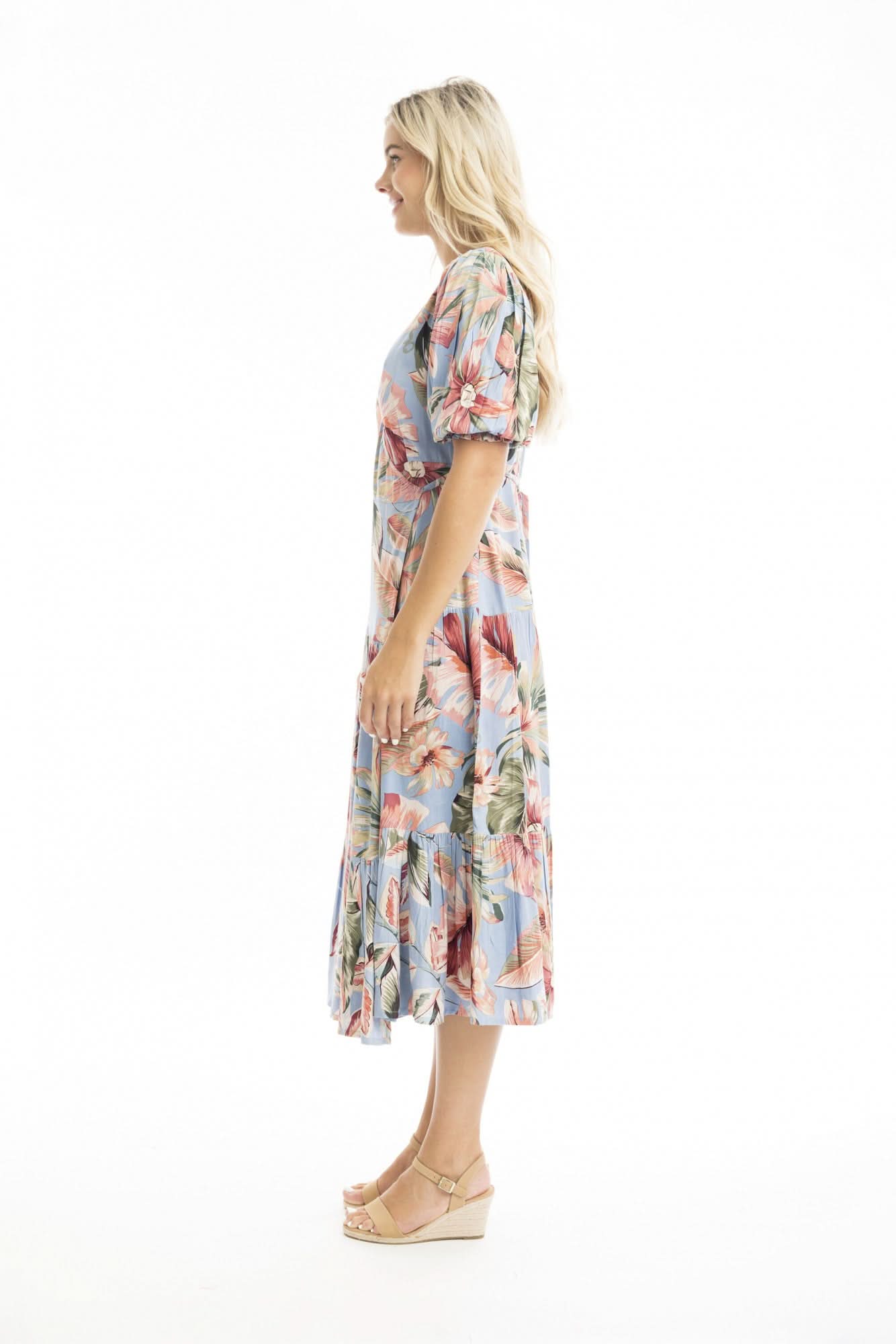 A woman models the Orientique Luzon Midi Crossover Dress—a light blue, floral-patterned summer dress with a v-neckline, short sleeves, and ruffled hem—paired with tan heeled sandals against a white background.