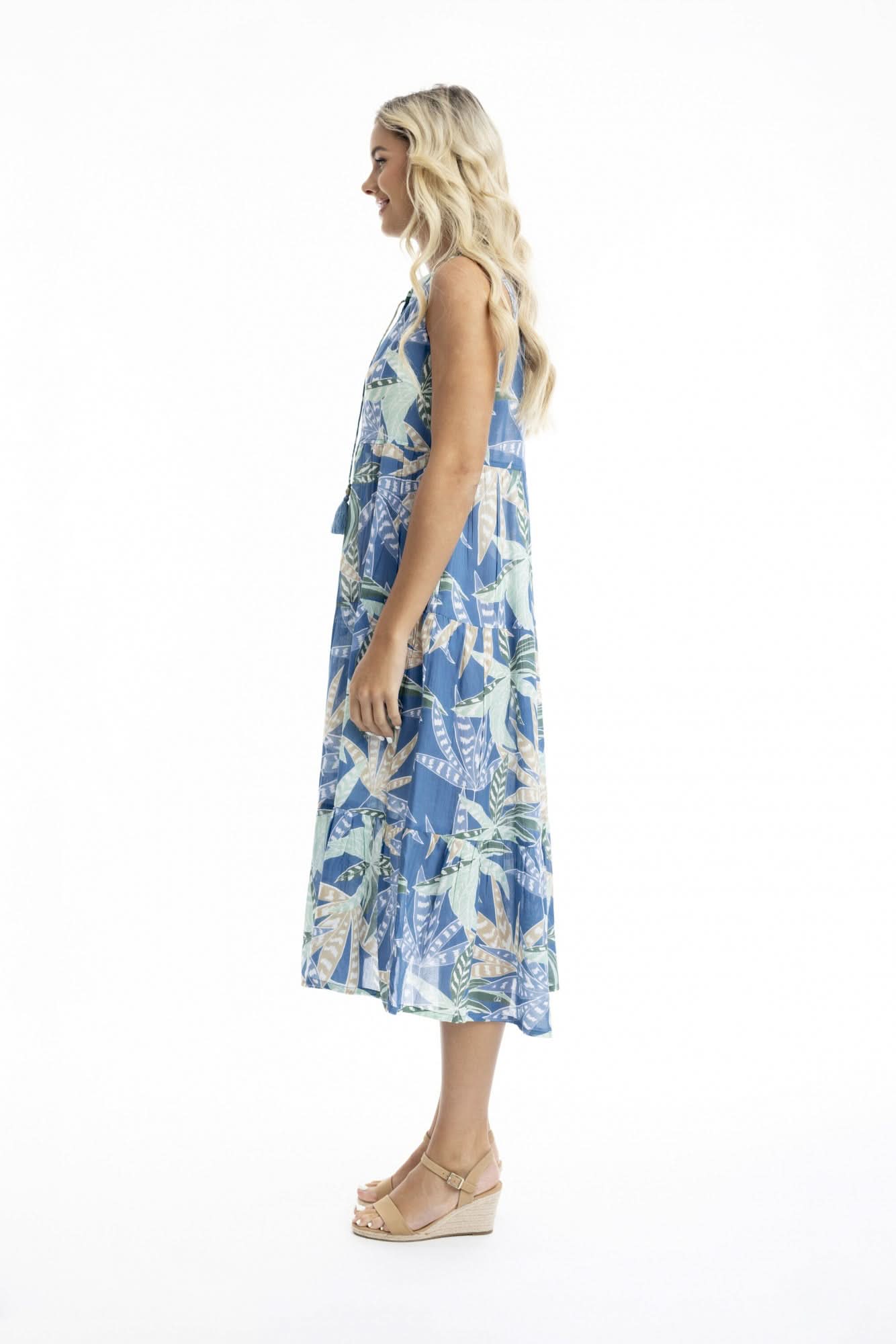 A woman poses against a white backdrop in the Orientique Fitzroy Sleeveless Boho Dress, featuring a blue tropical leaf print and v-neckline, paired with tan wedges—an ideal choice for summer events.