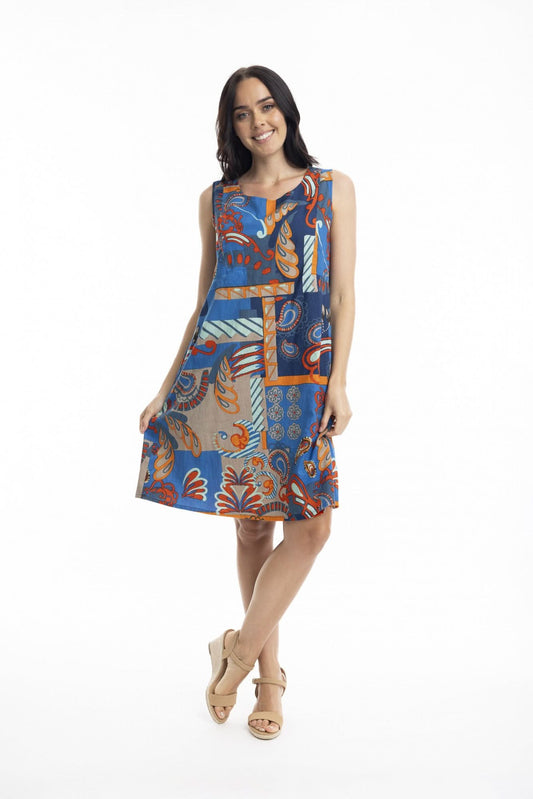 A woman stands smiling in the Orientique Camps Bay Sleeveless Reversible Dress, featuring a vibrant geometric and floral pattern, paired with beige high-heeled sandals against a white background—an ideal summer look.