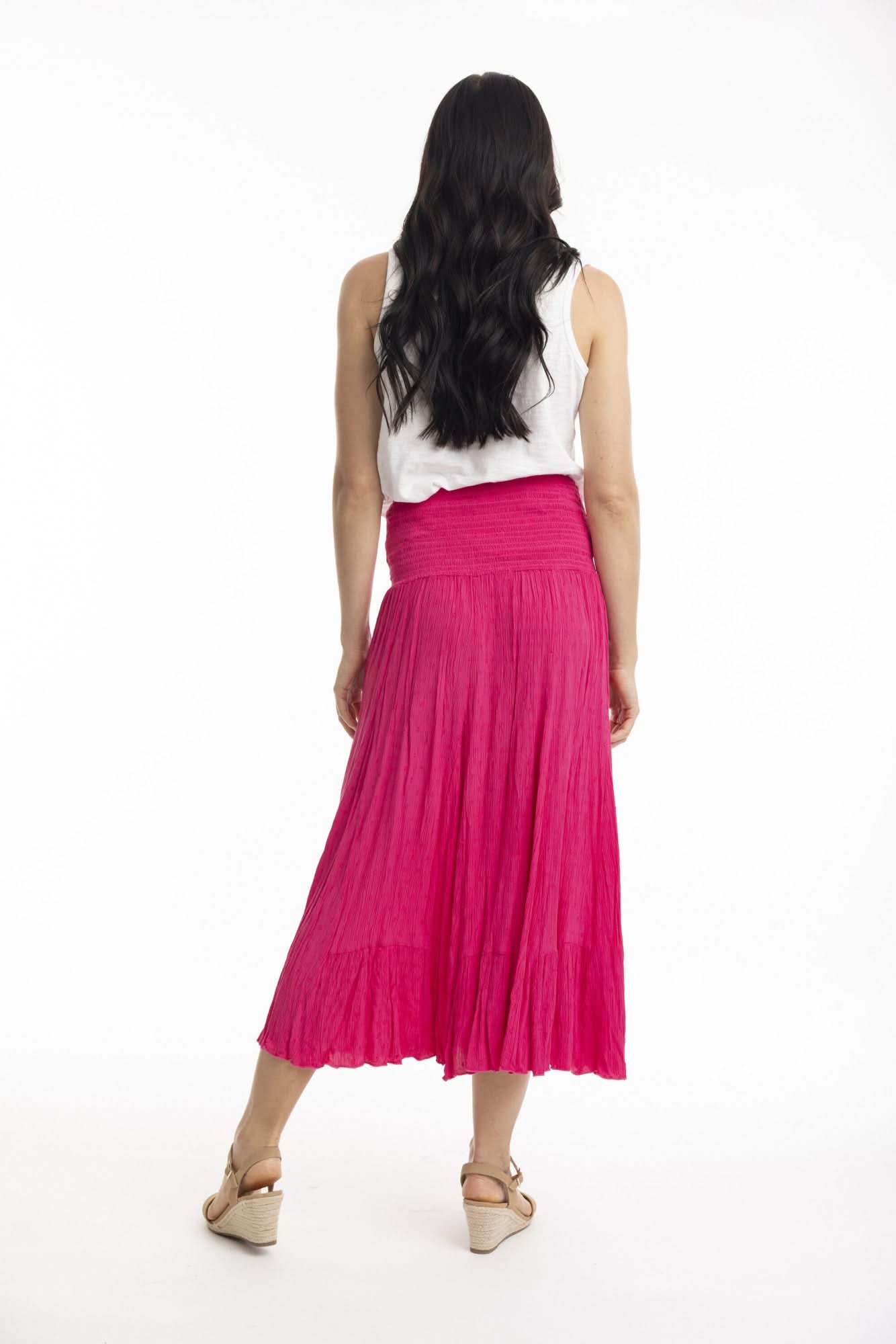A woman stands against a white background wearing the Orientique Essentials Dobby Skirt Dress— a sleeveless, navy blue, knee-length style — paired with beige sandals and holding the sides of her dress.