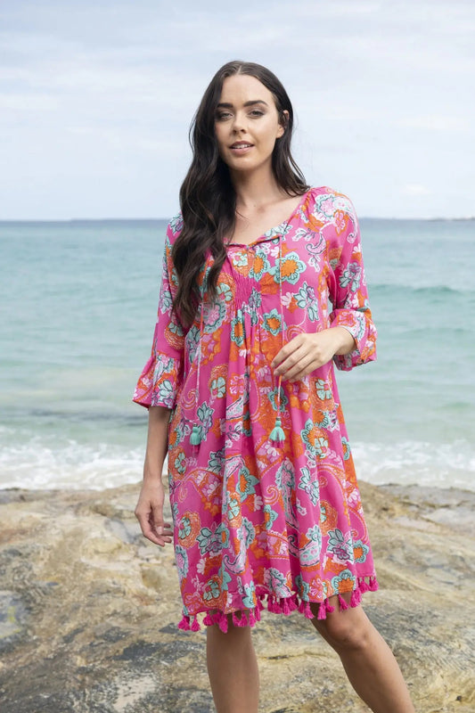 A woman in a floral pink Orientique Symi Dress Tassel standing on a rocky beach with the ocean in the background.