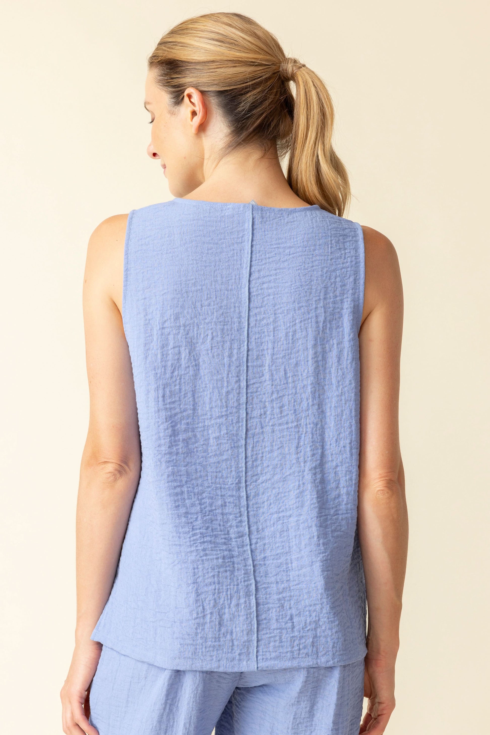 A woman smiles with her eyes closed, wearing the Habitat Pieces Tunic Tank and matching pants in sleeveless light blue—breathable fabric ensures comfort and style against a plain background.