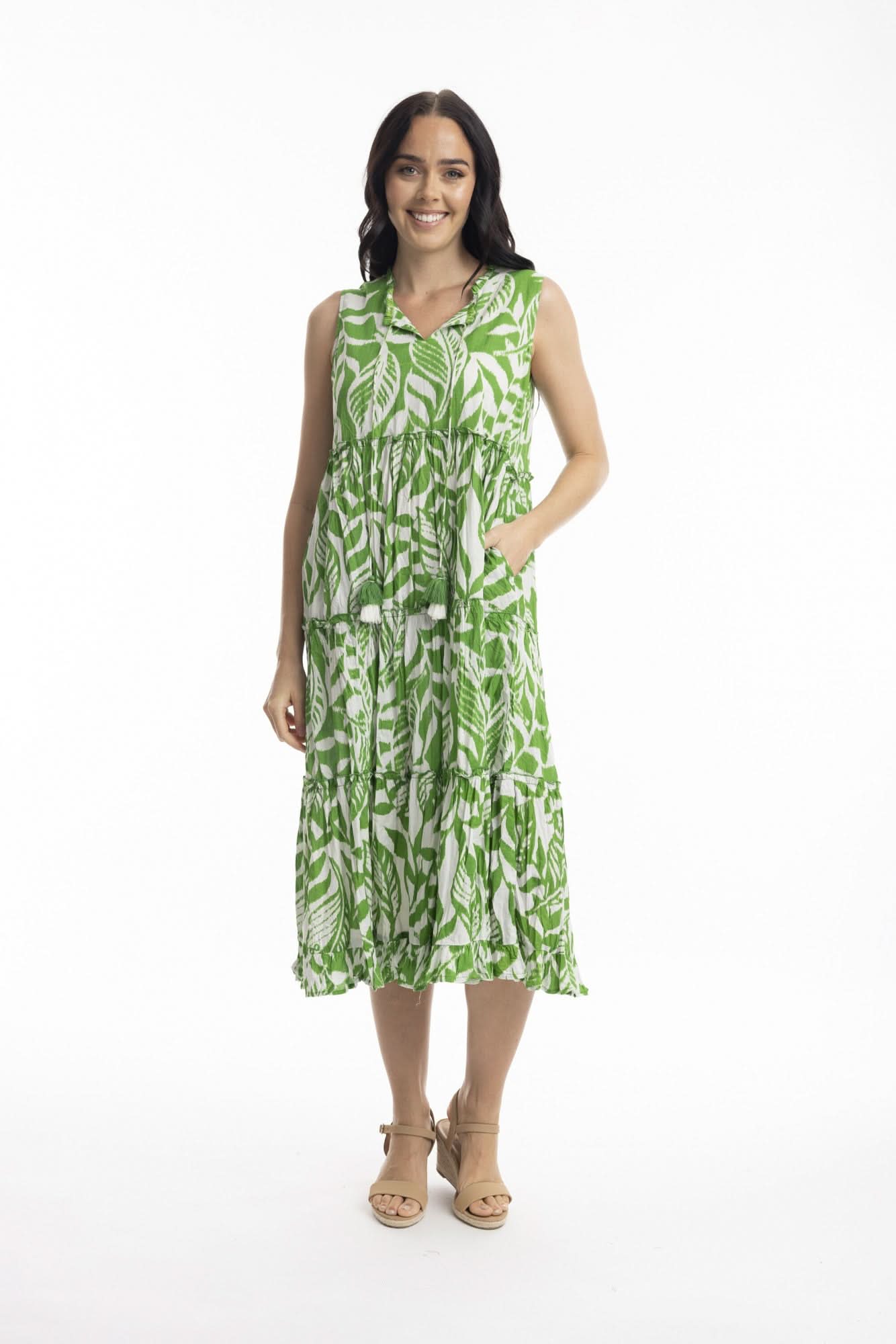 A woman smiles in the Orientique Hayman Green Sleeveless Boho Dress, featuring a green and white leaf pattern and tiered skirt, styled with beige heeled sandals—ideal for summer days against a plain white background.