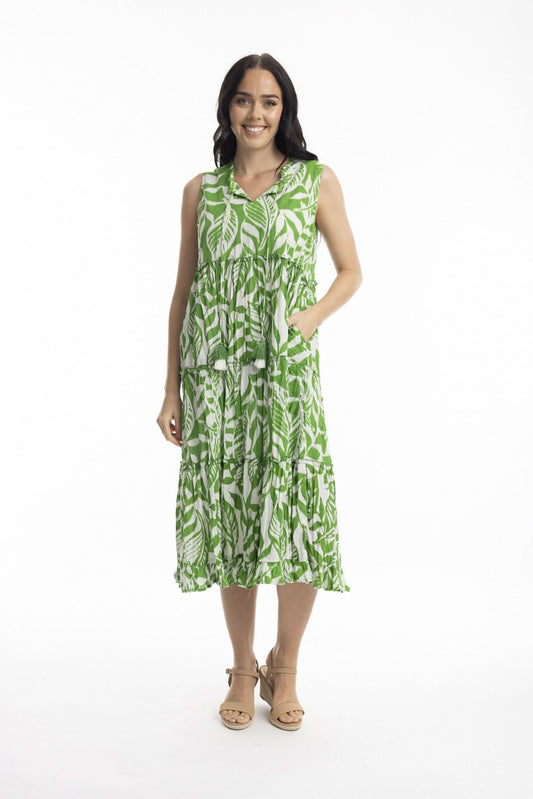 A woman smiles in the Orientique Hayman Green Sleeveless Boho Dress, featuring a green and white leaf pattern and tiered skirt, styled with beige heeled sandals—ideal for summer days against a plain white background.