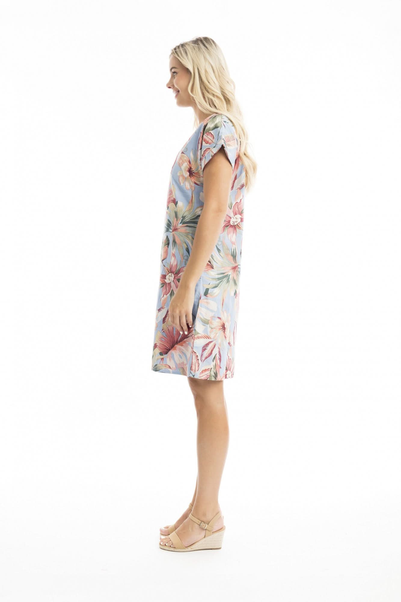 A woman stands against a white background wearing the Orientique Luzon Reversible Short Sleeve Dress, a floral knee-length piece for summer, styled with beige sandals and her hands in her pockets.