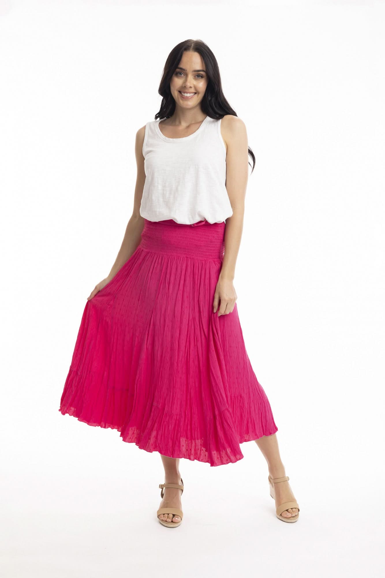 A woman stands against a white background wearing the Orientique Essentials Dobby Skirt Dress— a sleeveless, navy blue, knee-length style — paired with beige sandals and holding the sides of her dress.