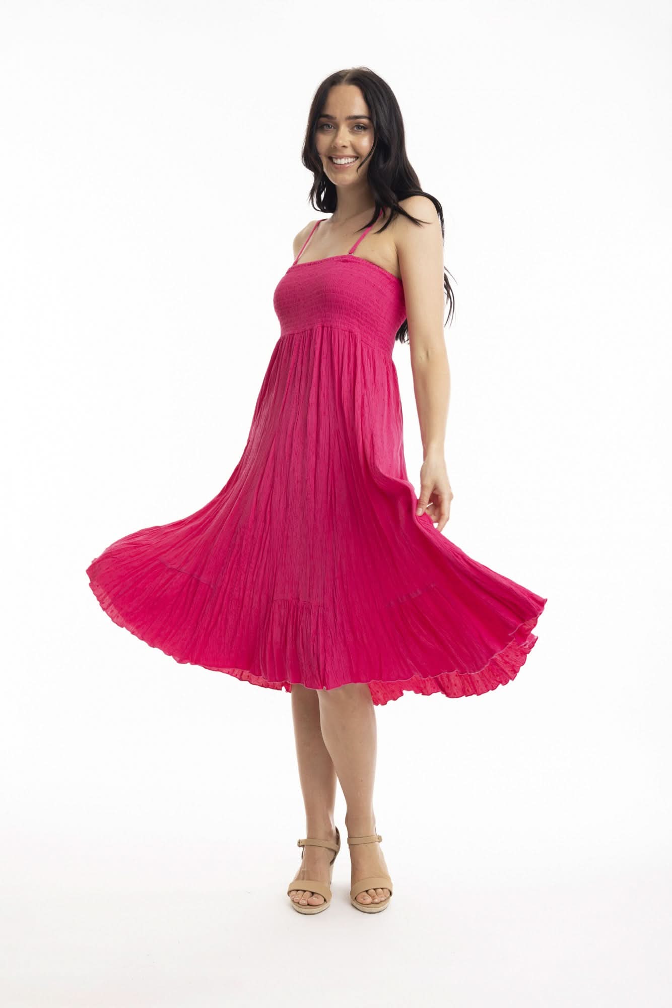 A woman stands against a white background wearing the Orientique Essentials Dobby Skirt Dress— a sleeveless, navy blue, knee-length style — paired with beige sandals and holding the sides of her dress.
