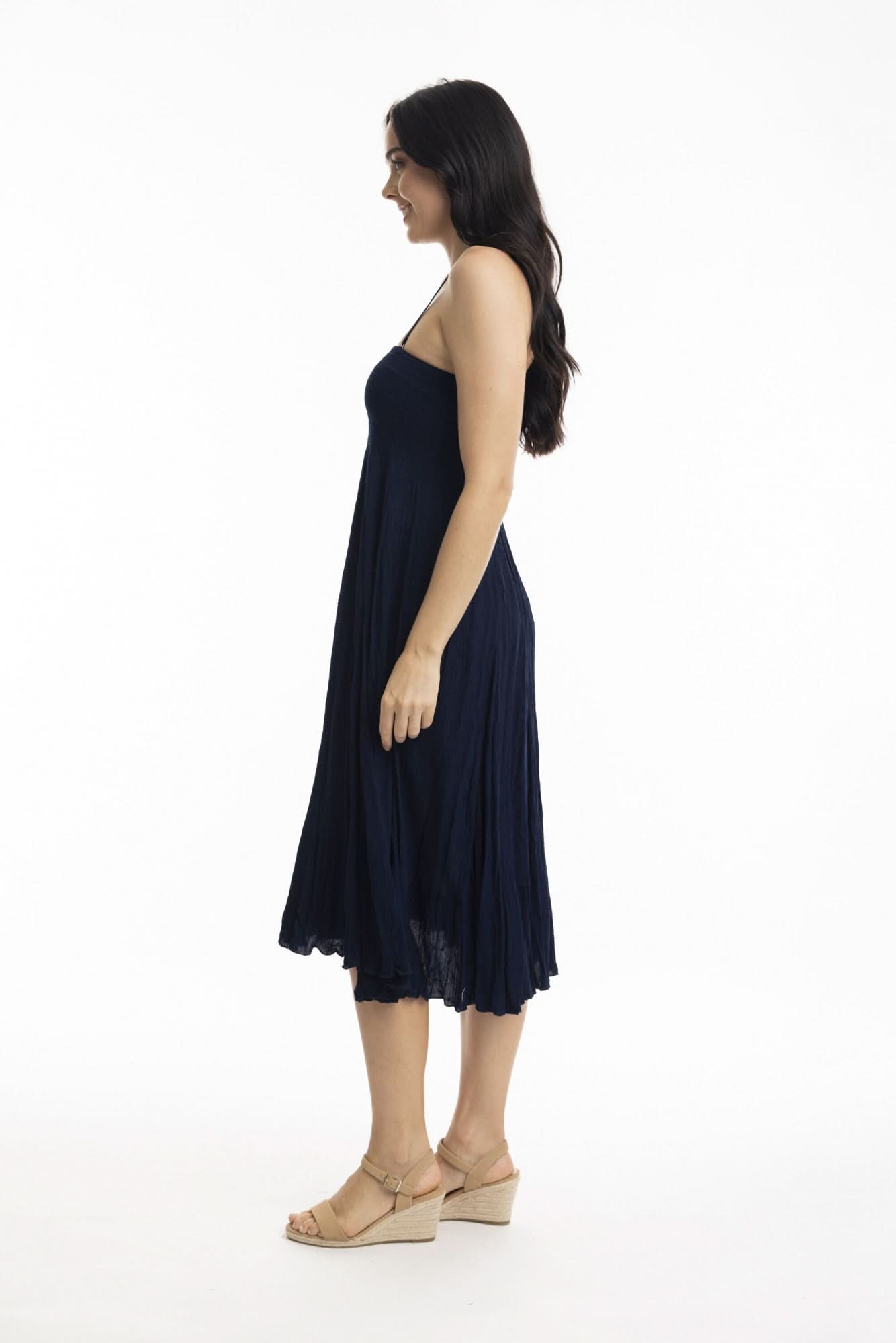 A woman stands against a white background wearing the Orientique Essentials Dobby Skirt Dress— a sleeveless, navy blue, knee-length style — paired with beige sandals and holding the sides of her dress.
