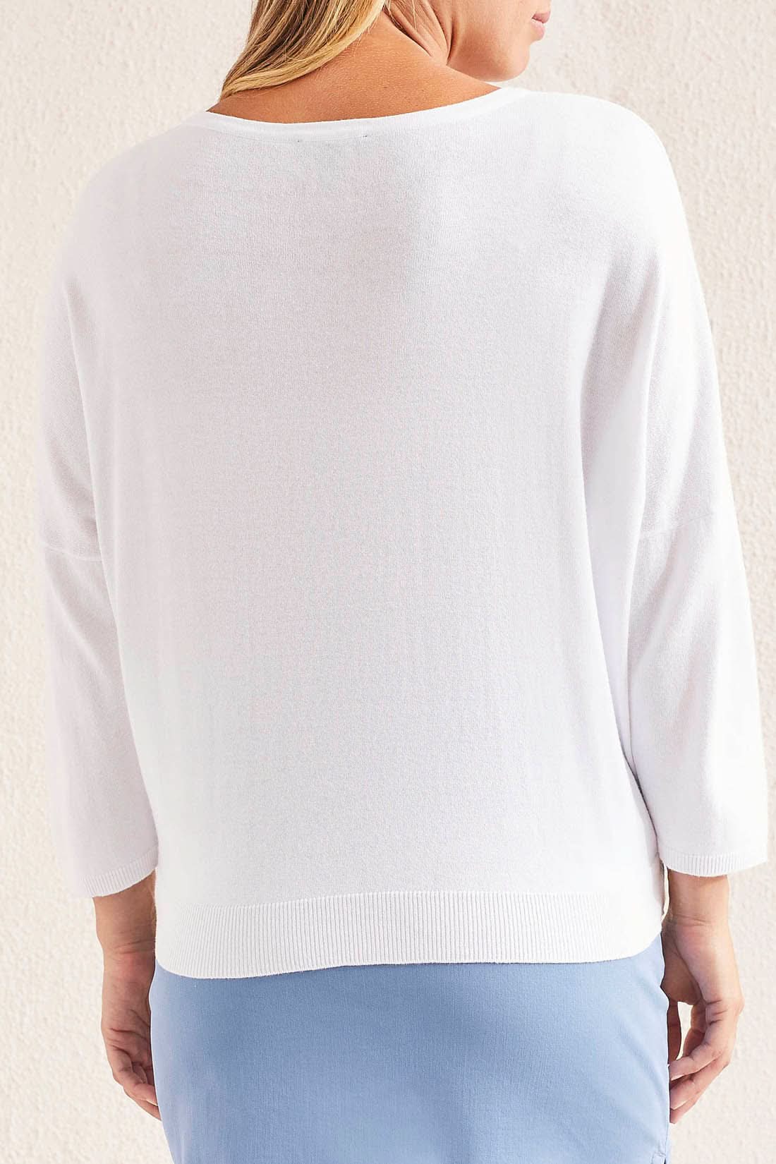 Woman wearing a cozy white Tribal sweater with blue abstract design, a light blue skirt, and white sneakers, standing against a neutral background.