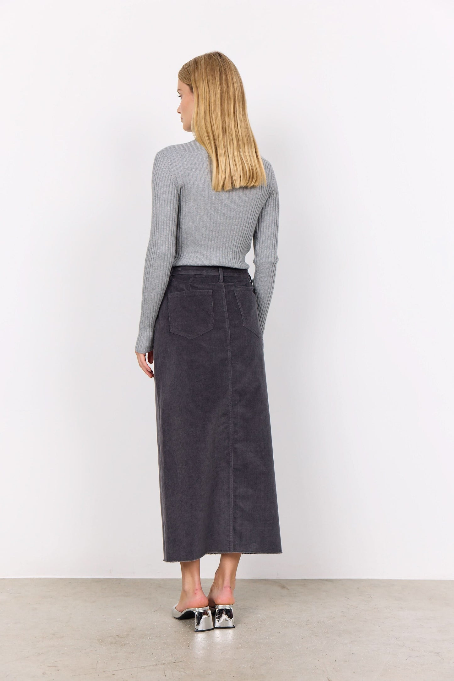 Person wearing a gray corduroy Soya Concepts IVALOU 2 Long Skirt with a front slit and a gray long-sleeve top, standing on a concrete floor in silver open-toe heeled sandals, exuding effortless style.
