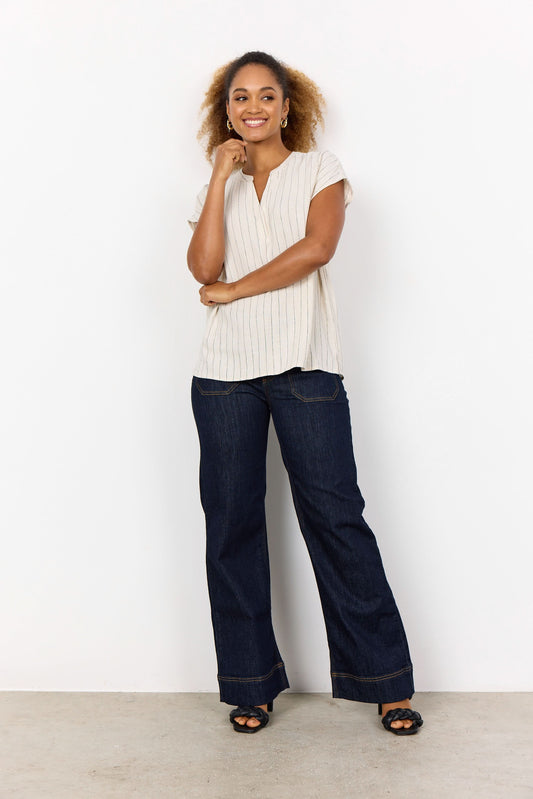 A woman stands against a white background, smiling and touching her hair. She is wearing the Soya Concepts PAULA 1 Top, a light ECOVERO Viscose blouse with thin vertical stripes, paired with dark jeans.
