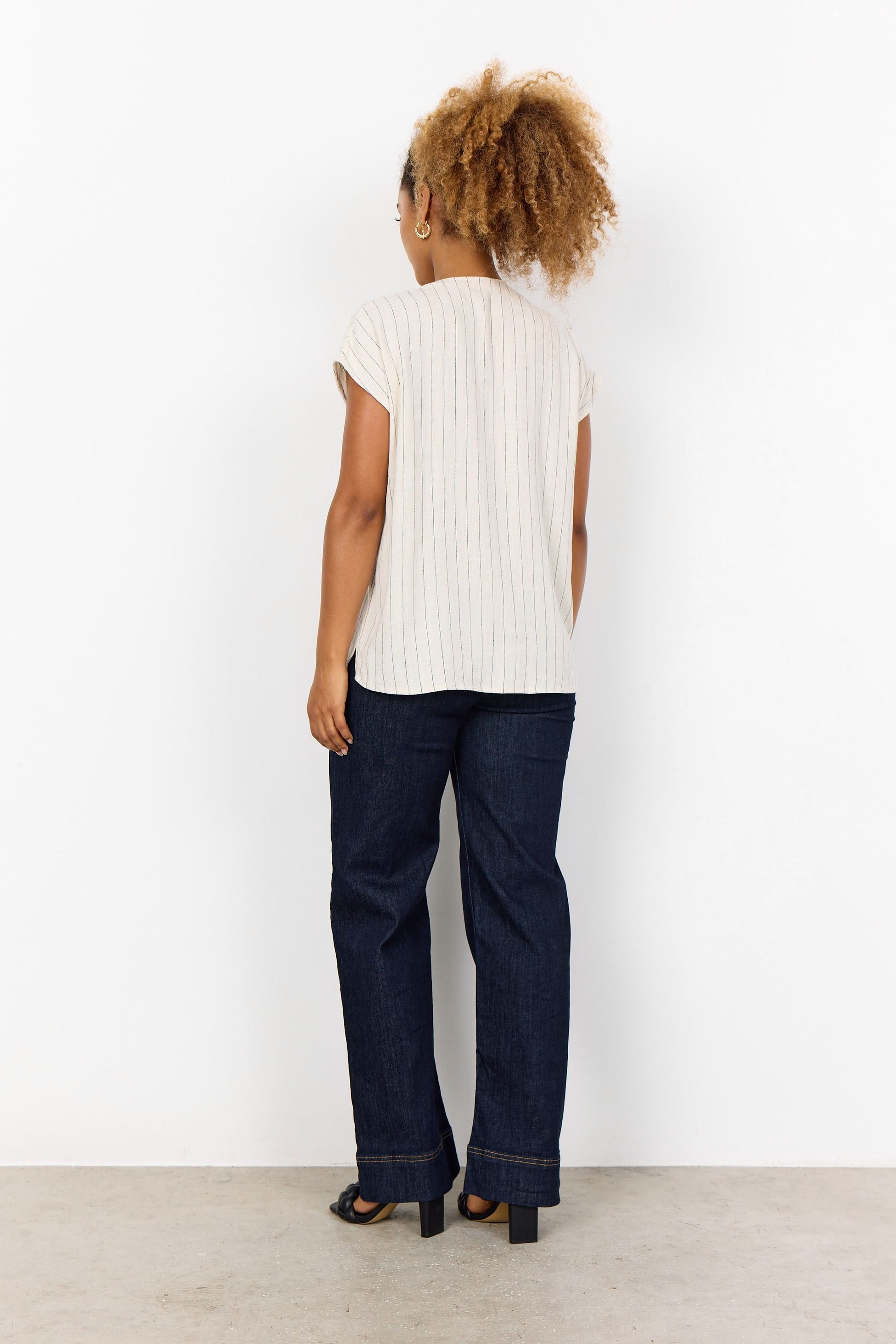 A woman stands against a white background, smiling and touching her hair. She is wearing the Soya Concepts PAULA 1 Top, a light ECOVERO Viscose blouse with thin vertical stripes, paired with dark jeans.