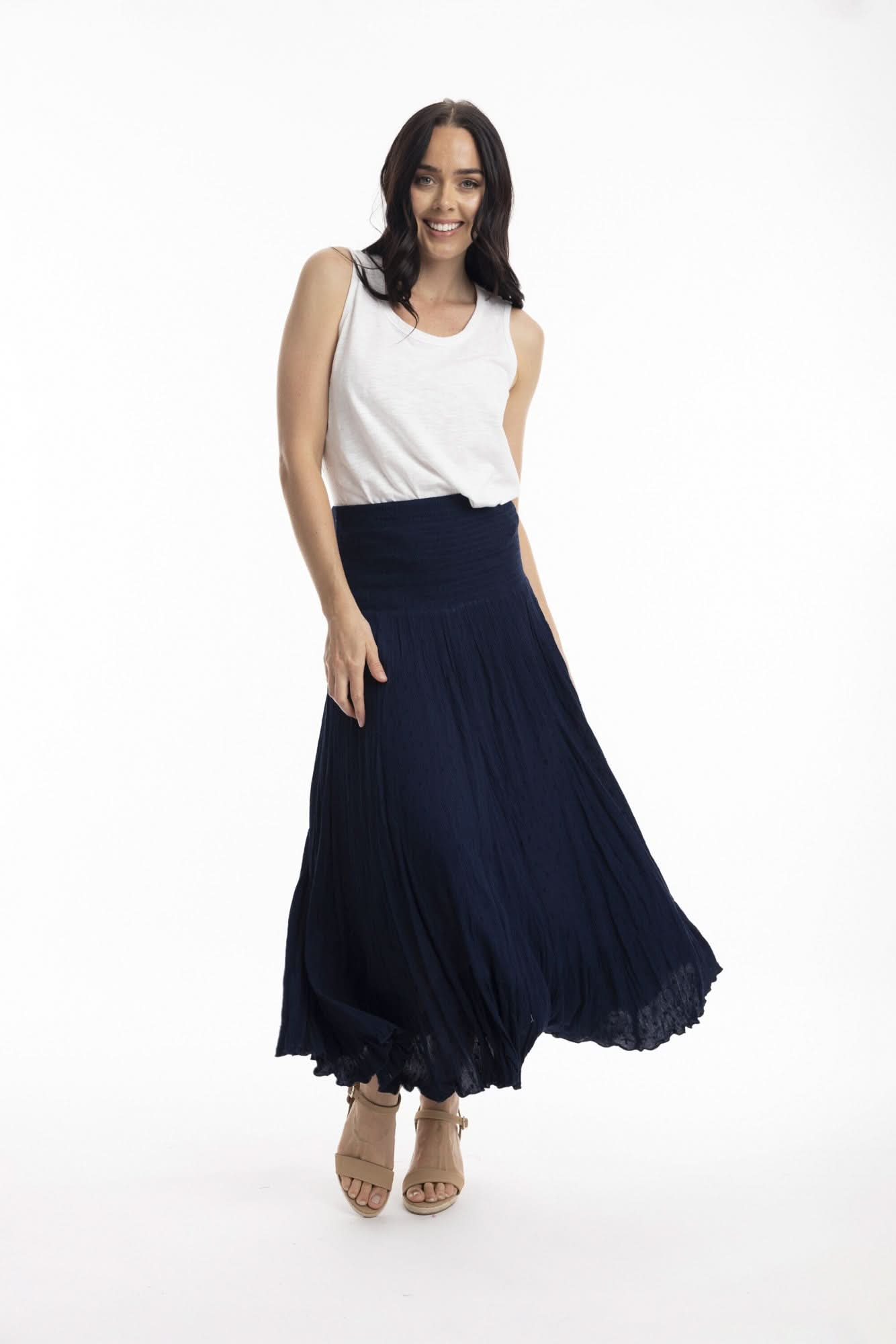 A woman stands against a white background wearing the Orientique Essentials Dobby Skirt Dress— a sleeveless, navy blue, knee-length style — paired with beige sandals and holding the sides of her dress.