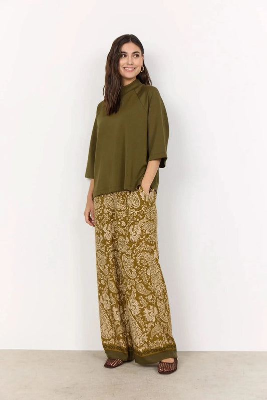 A woman stands against a plain light background wearing the Soya Concepts Short Sleeve Knit in dark olive, paired with brown paisley-patterned pants.