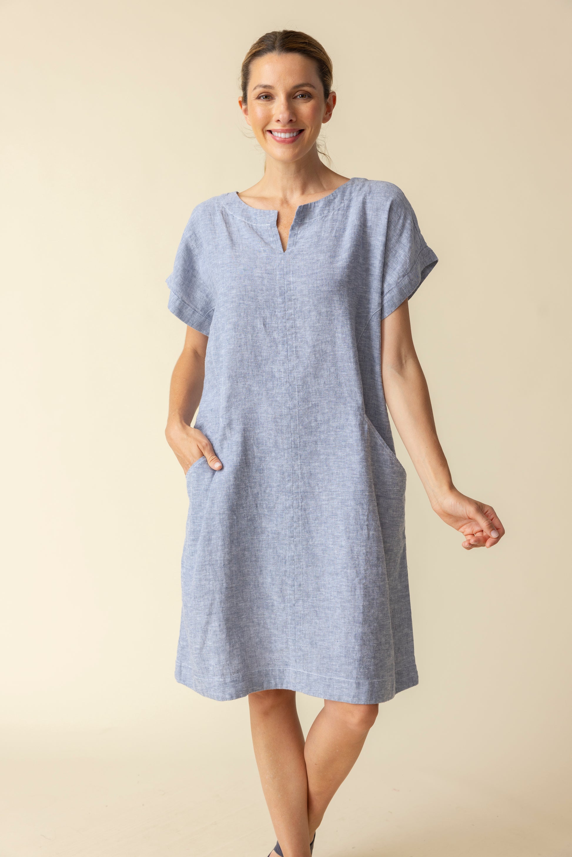 A woman wearing the Habitat Bethany Dress with Belt in light blue, featuring short sleeves, knee-length hem, tie belt, and pockets, stands against a plain beige background.