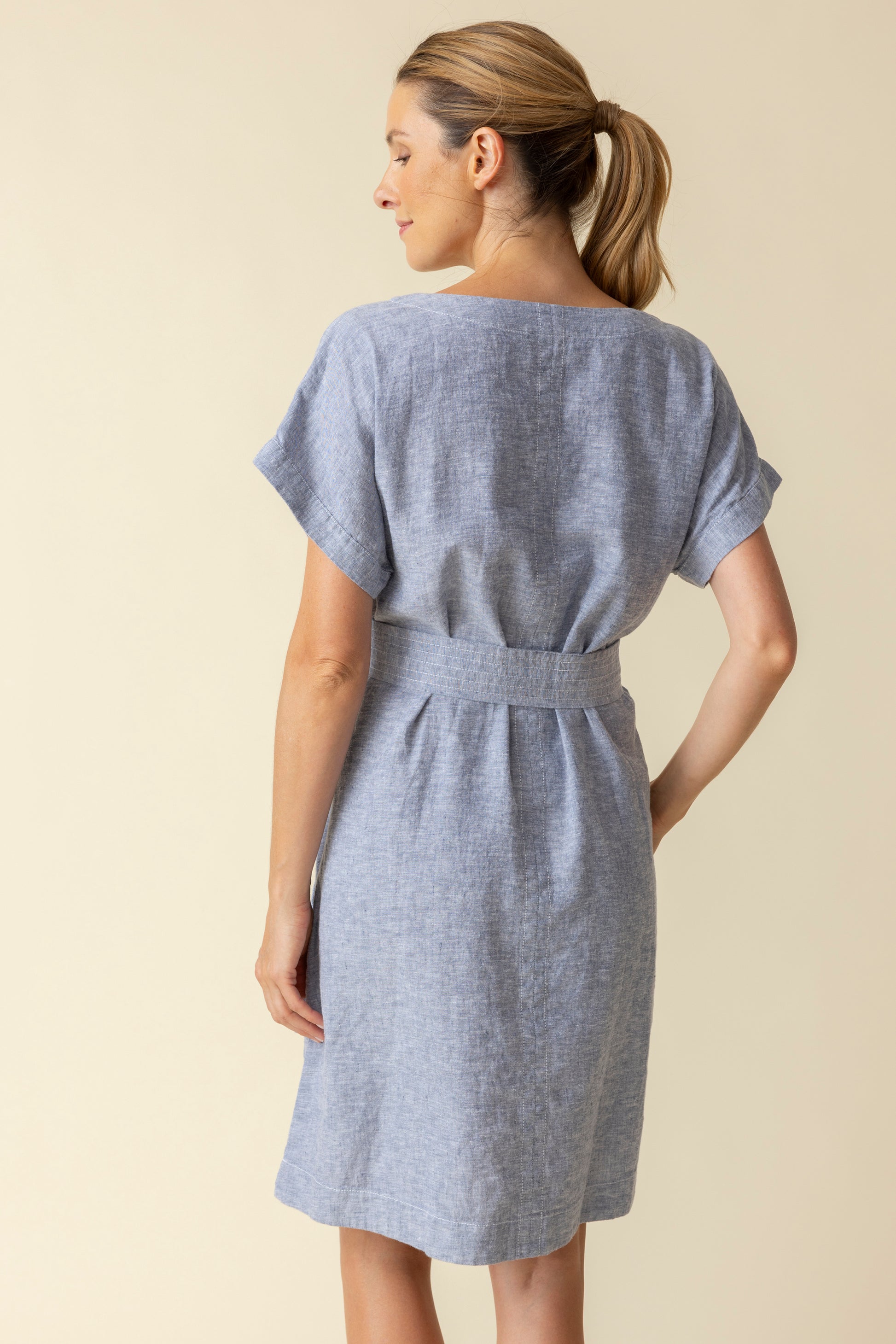 A woman wearing the Habitat Bethany Dress with Belt in light blue, featuring short sleeves, knee-length hem, tie belt, and pockets, stands against a plain beige background.
