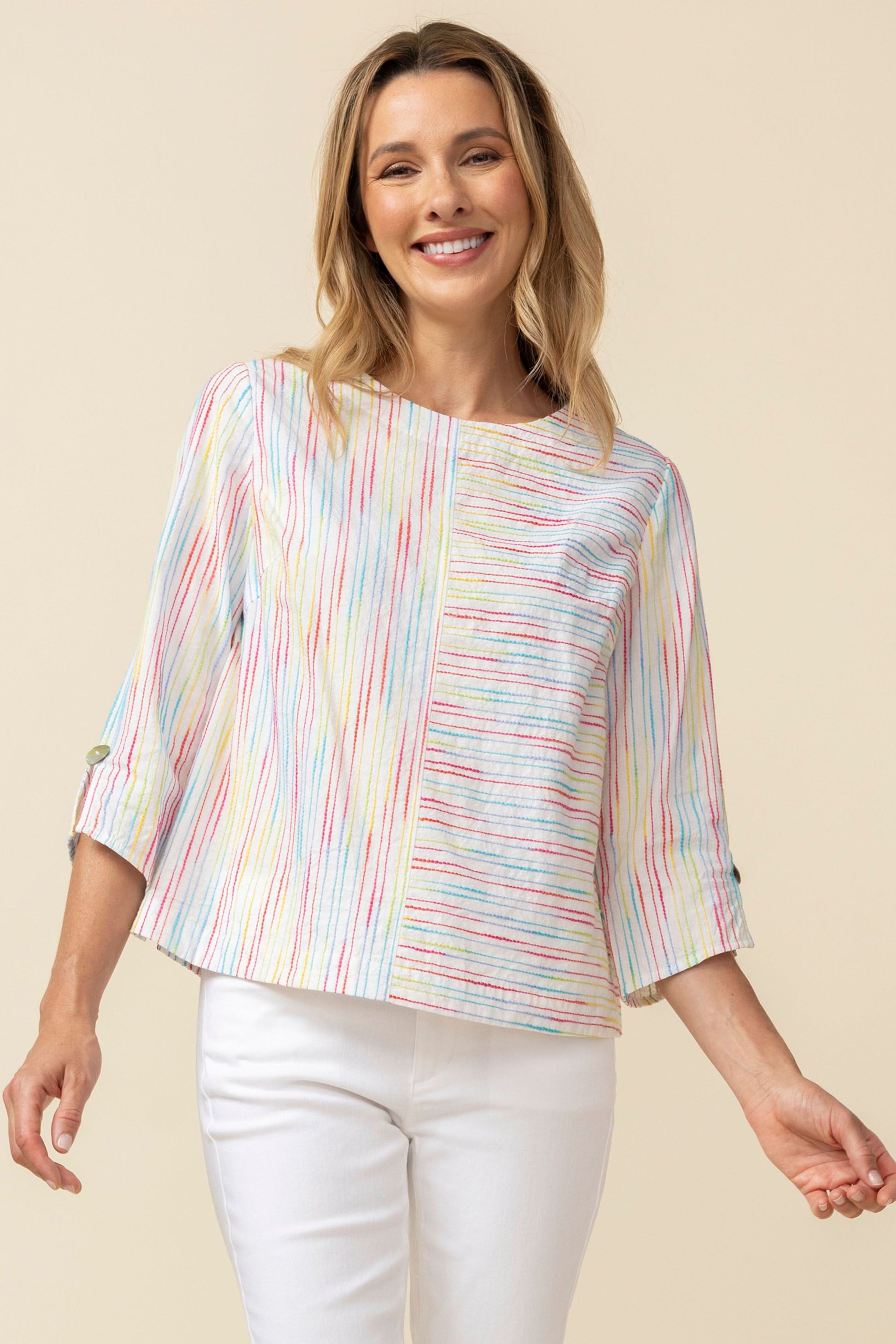 A woman smiles while wearing the Habitat Carousel Pullover, featuring a multi-color design and 3/4 sleeves, paired with white pants against a plain beige background.