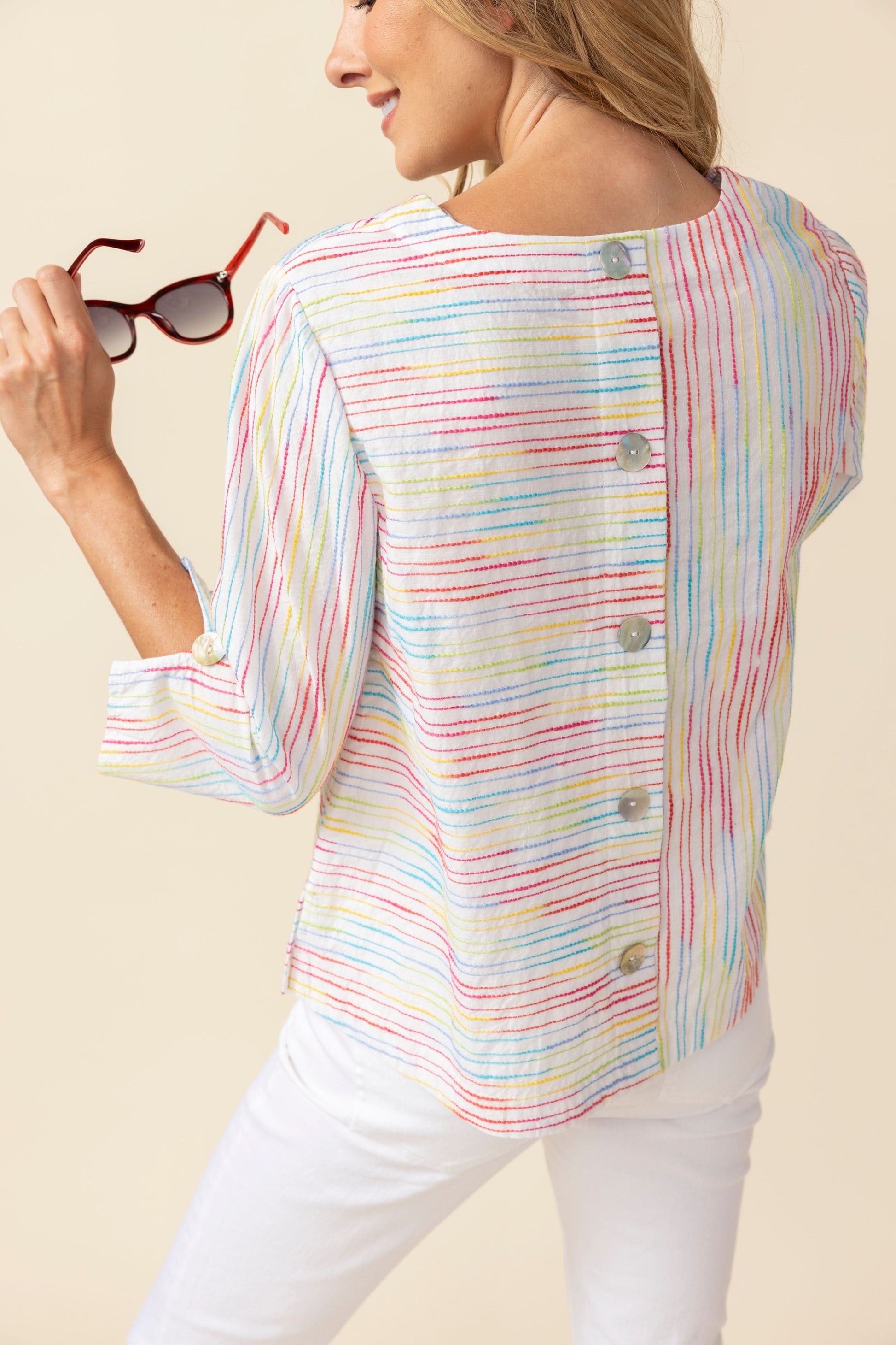 A woman smiles while wearing the Habitat Carousel Pullover, featuring a multi-color design and 3/4 sleeves, paired with white pants against a plain beige background.