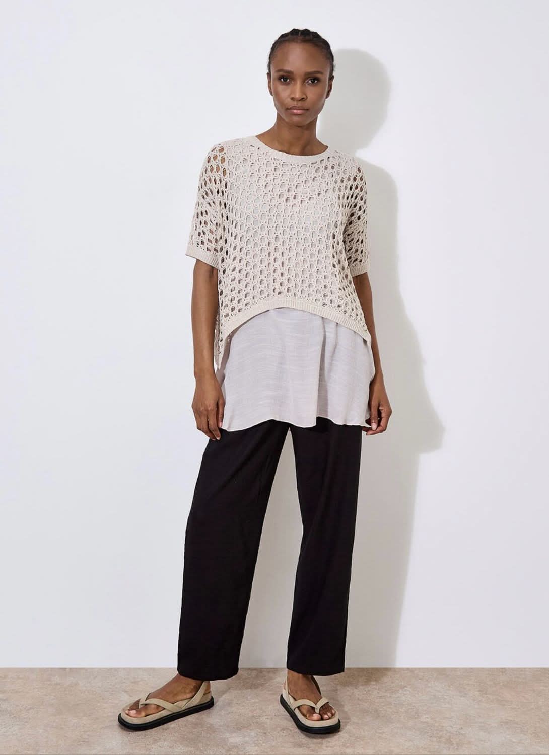 Against a plain, light backdrop, the individual wears Apricot's Knit Mesh Slub Tunic over a white blouse. This textured cream-crochet top features short sleeves and adds elegance as they gaze toward the camera.