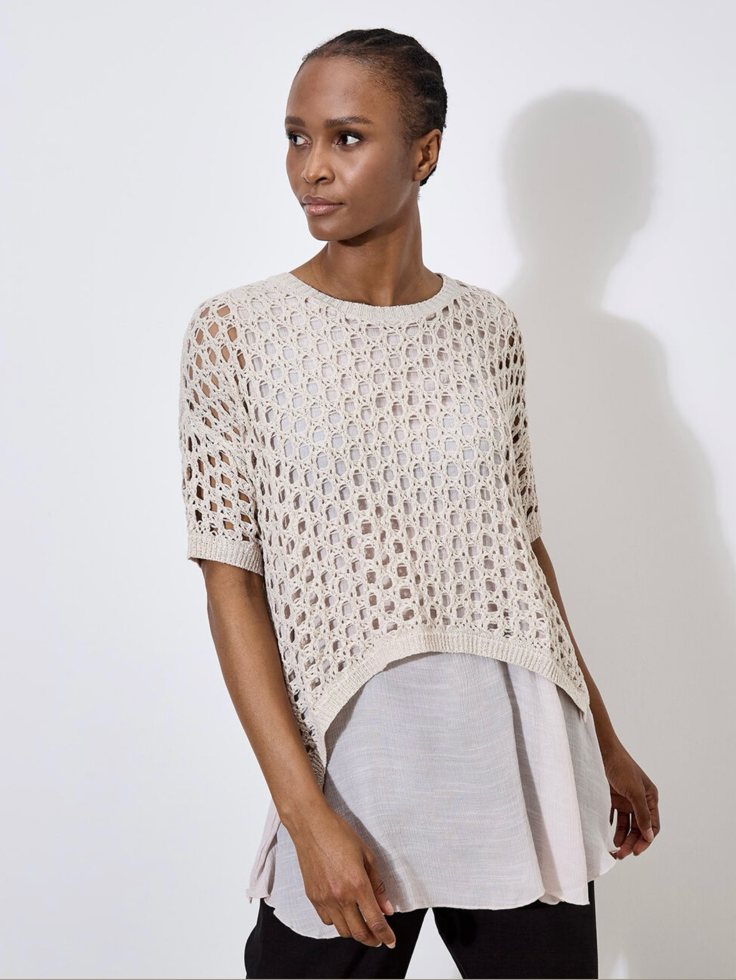 Against a plain, light backdrop, the individual wears Apricot's Knit Mesh Slub Tunic over a white blouse. This textured cream-crochet top features short sleeves and adds elegance as they gaze toward the camera.