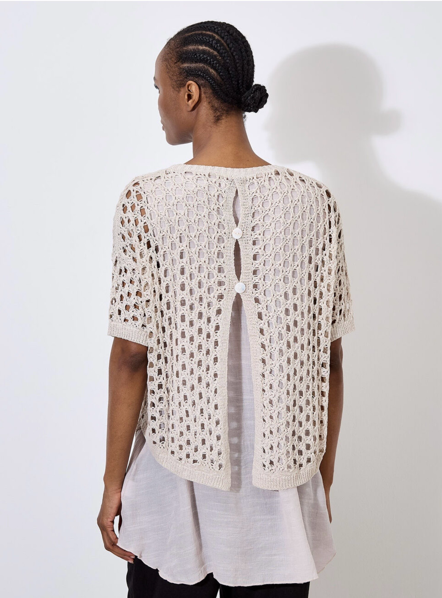 Against a plain, light backdrop, the individual wears Apricot's Knit Mesh Slub Tunic over a white blouse. This textured cream-crochet top features short sleeves and adds elegance as they gaze toward the camera.