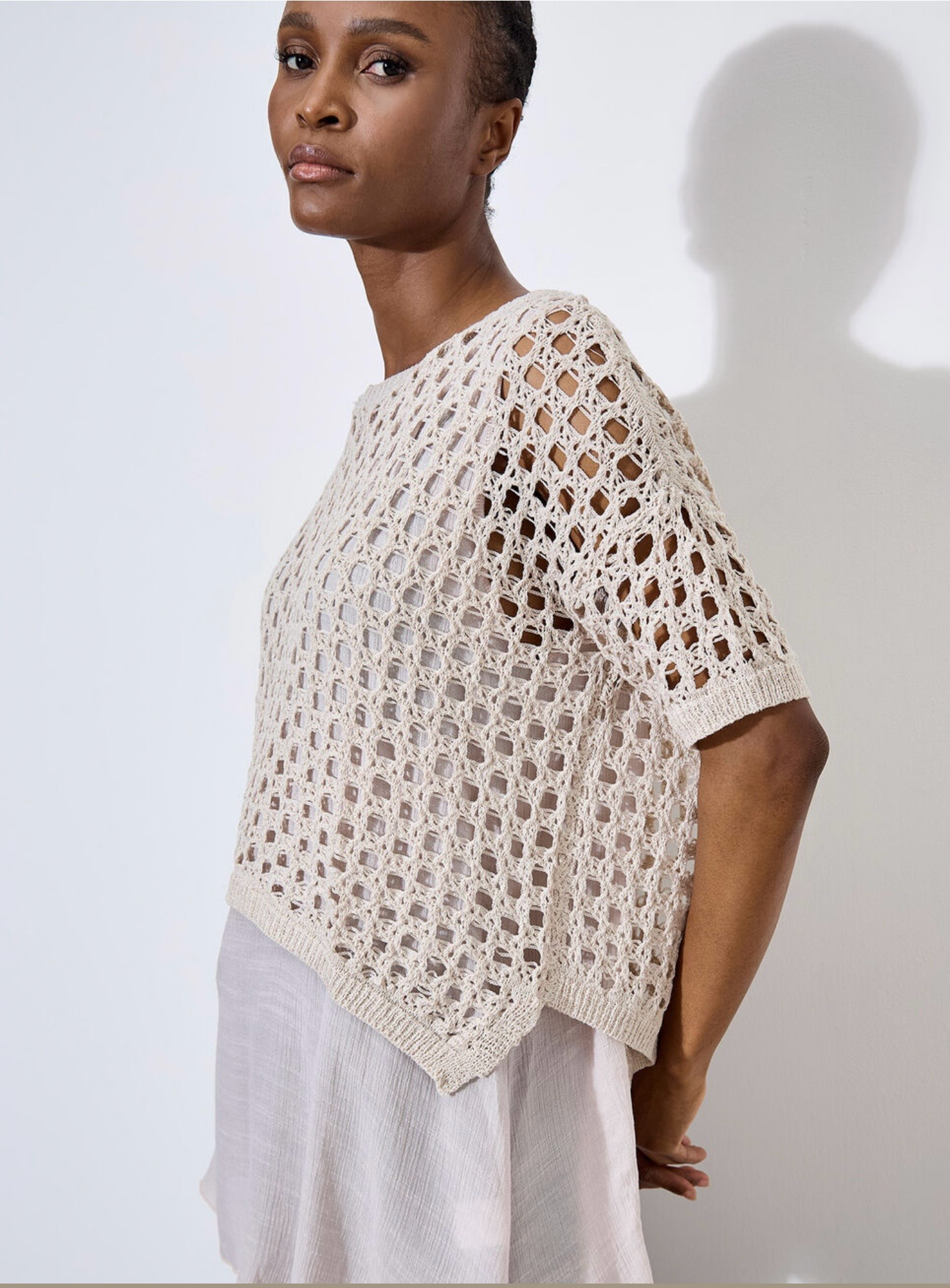 Against a plain, light backdrop, the individual wears Apricot's Knit Mesh Slub Tunic over a white blouse. This textured cream-crochet top features short sleeves and adds elegance as they gaze toward the camera.