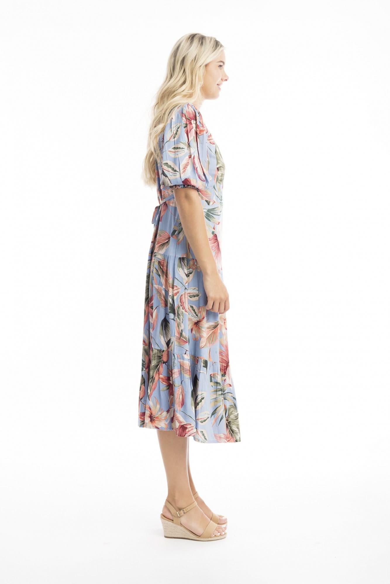 A woman models the Orientique Luzon Midi Crossover Dress—a light blue, floral-patterned summer dress with a v-neckline, short sleeves, and ruffled hem—paired with tan heeled sandals against a white background.
