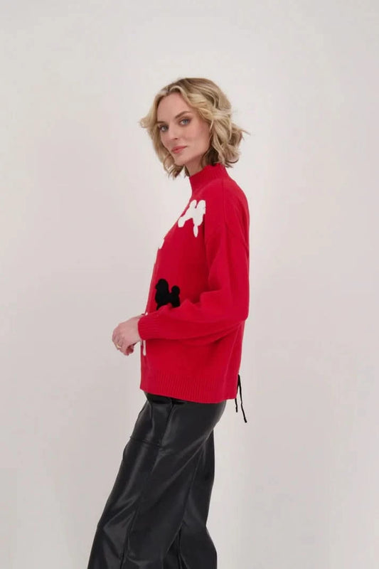 Against a plain backdrop, a poodle enthusiast exhibits their fashion sense by wearing the Poodle Turtleneck Sweater With Back-Tie Detail from Gabby Isabella—a striking red sweater embellished with white and black poodle motifs—and pairs it seamlessly with black pants.